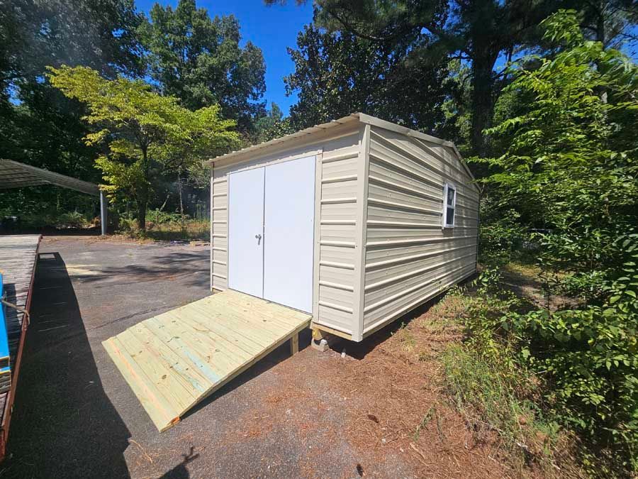 Tan metal shed with white double doors, a wooden ramp, and a small window.