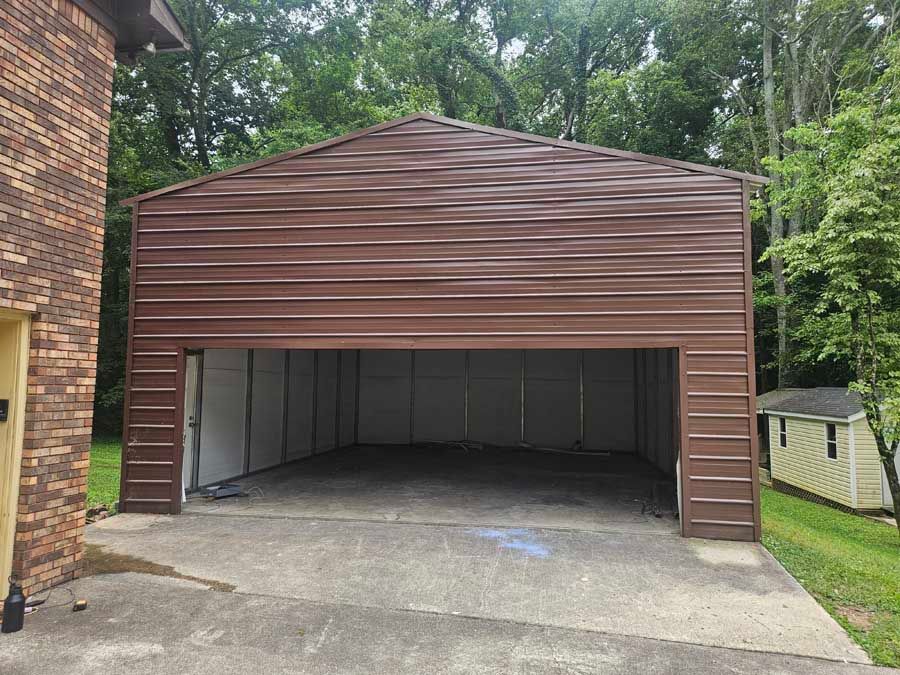 Brown metal garage with open door, concrete pad, next to a brick building, wooded area in background.