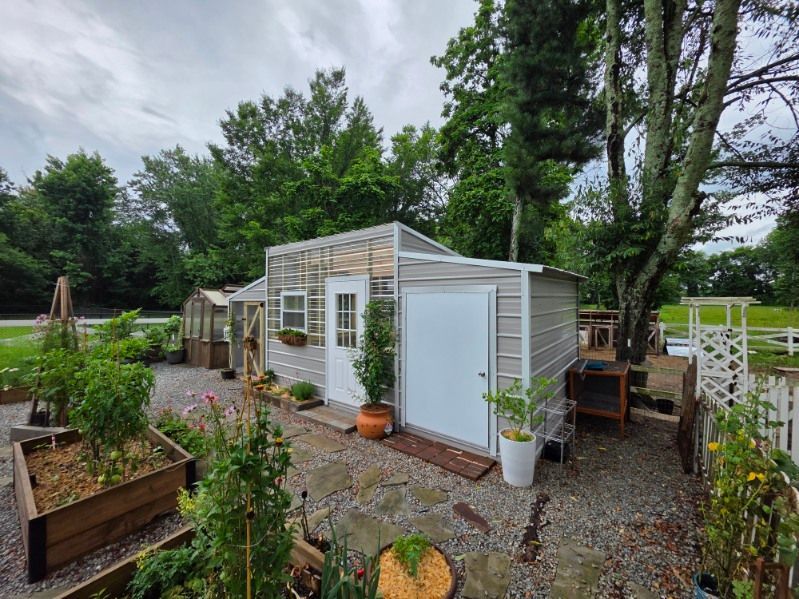 White garden shed with pergola, surrounded by potted plants and gravel path in a lush backyard