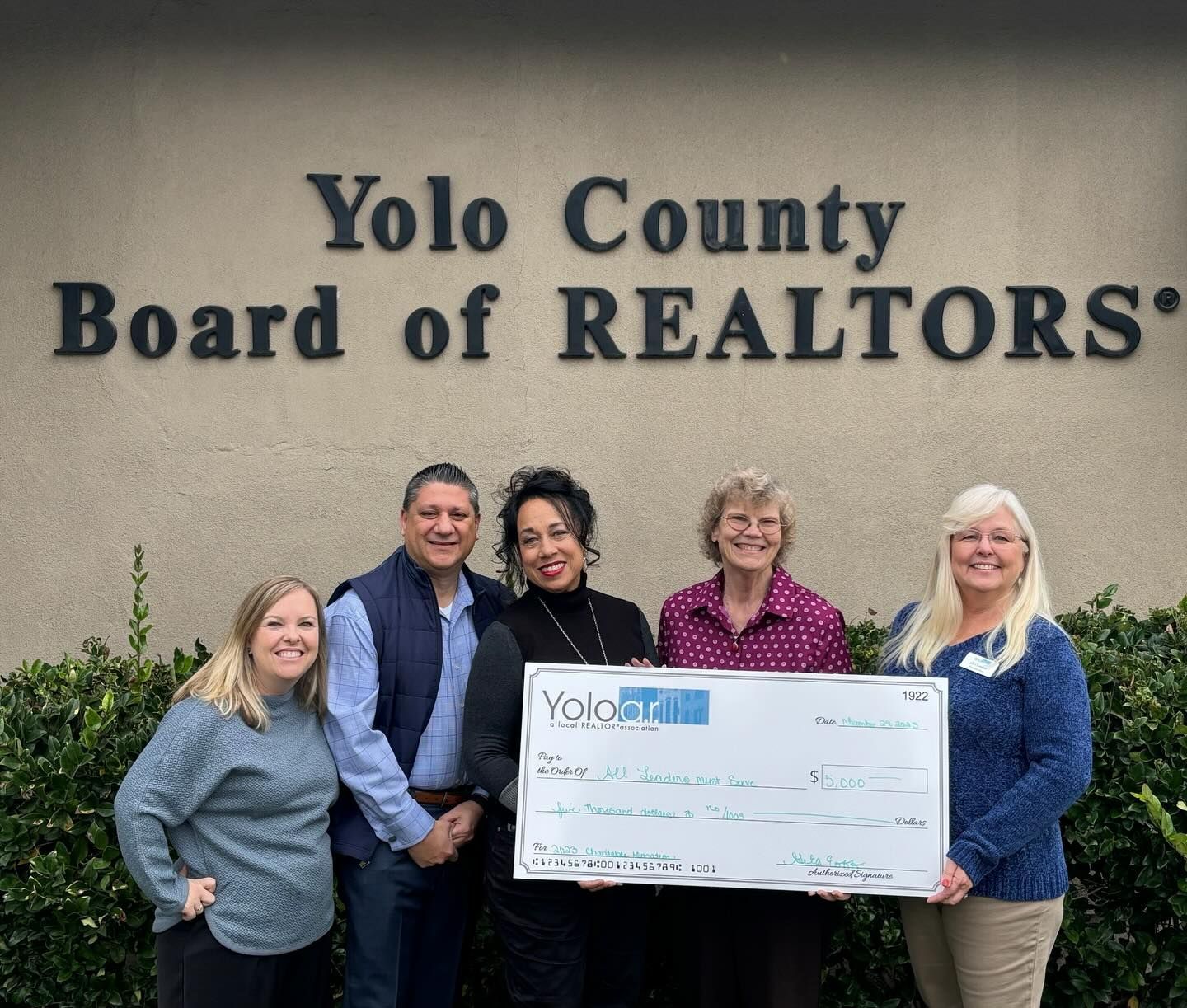 Five people stand smiling in front of a Yolo County Board of REALTORS sign, holding a large ceremonial donation check.