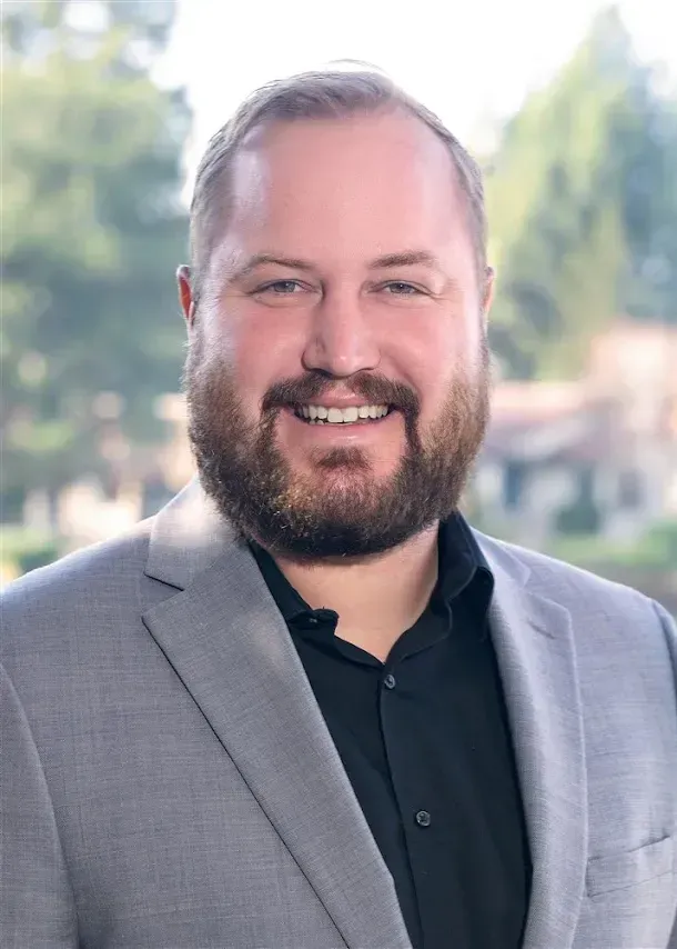 Professional headshot of a person with a beard, wearing a light gray blazer and black shirt, outdoors with blurred trees.