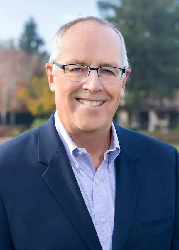 A smiling person wearing glasses, a light-colored button-down shirt, and a dark blazer, posed outdoors in front of trees.