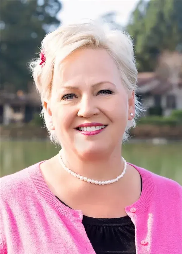 A smiling person with short blonde hair and a small red hair accessory, wearing a pink cardigan and a pearl necklace.