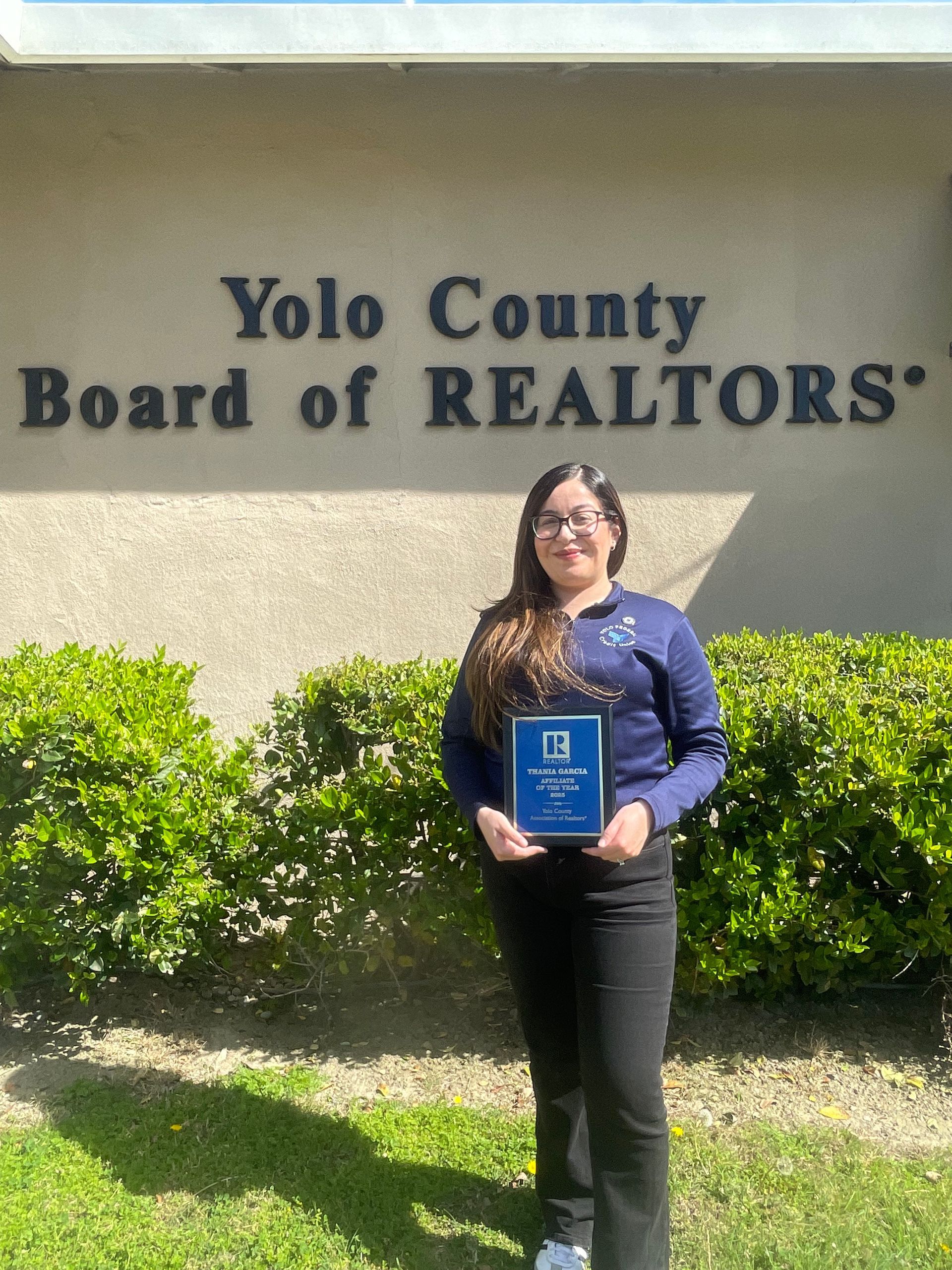 A person holds a blue award plaque in front of the Yolo County Board of Realtors building, standing before green bushes.
