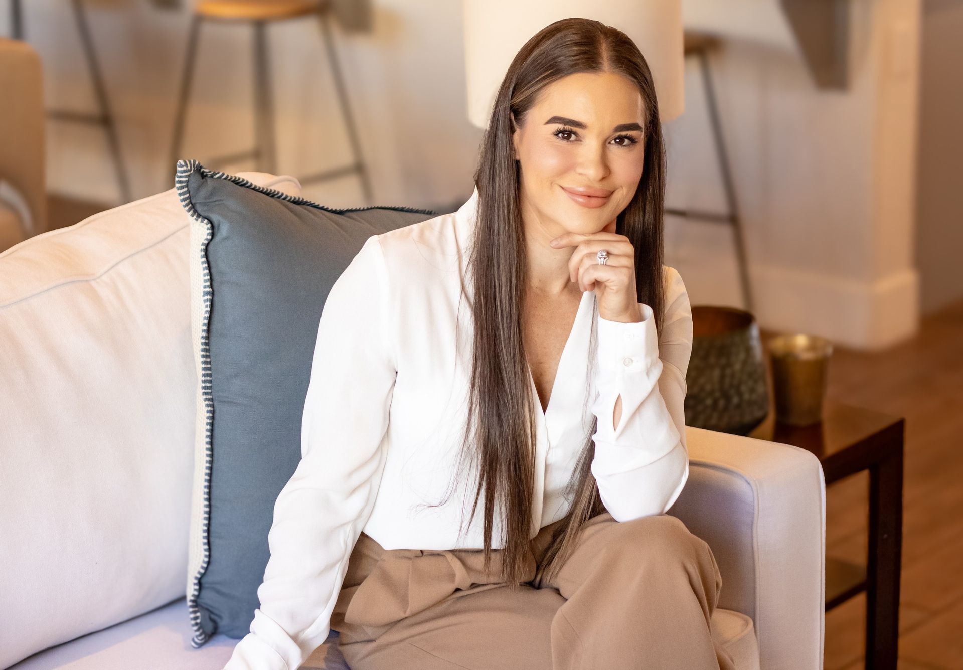 A person with long brown hair, wearing a white shirt and tan trousers, poses on a white couch, resting chin on a hand.