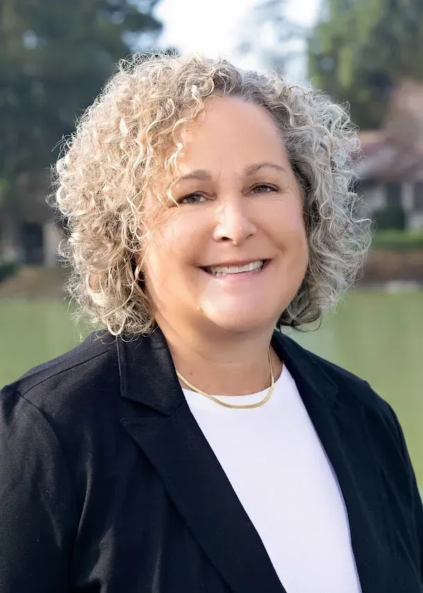 A smiling person with curly, shoulder-length blonde hair wearing a black blazer over a white shirt.