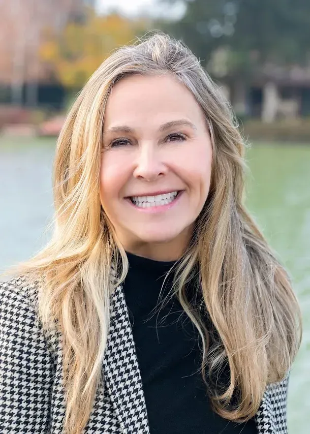 A person with long blonde hair smiling, wearing a black turtleneck and a black-and-white houndstooth blazer outdoors.