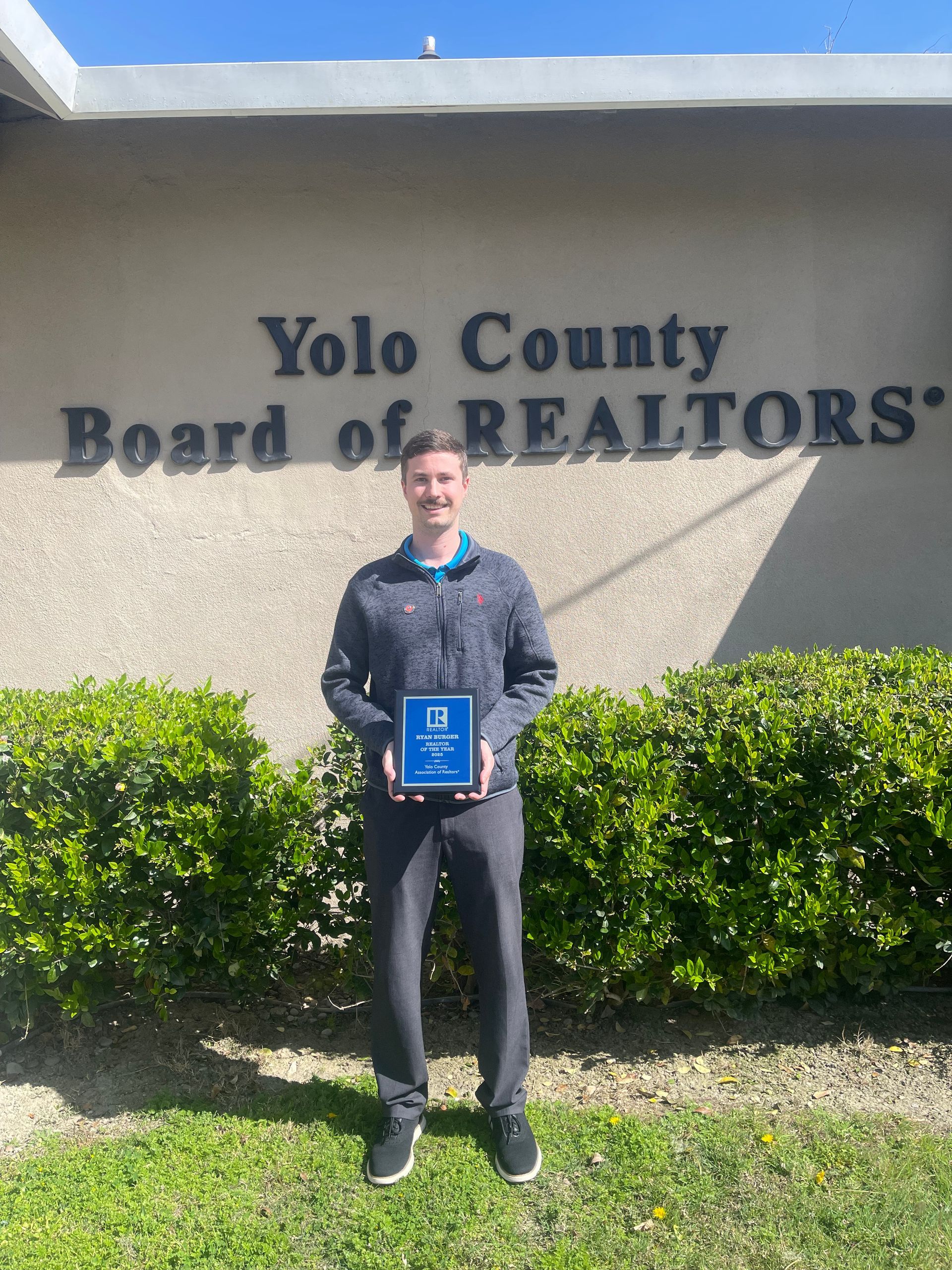A person holding a small award plaque in front of the Yolo County Board of Realtors building.
