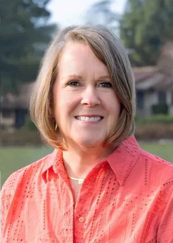 A person with shoulder-length brown hair, smiling, wearing a coral, short-sleeved, button-down shirt with a subtle pattern.