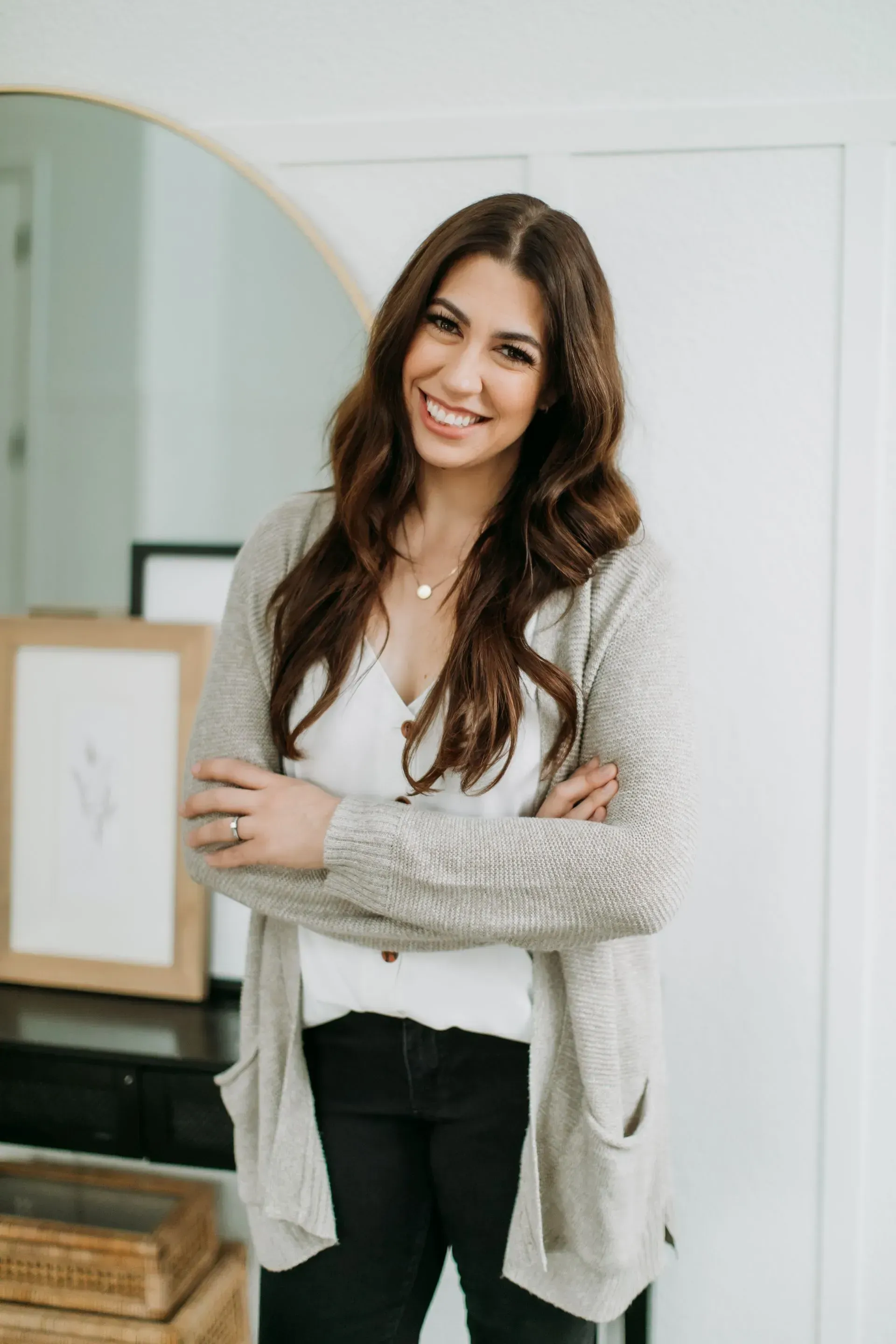 A smiling person with long brown hair, wearing a white shirt and beige cardigan, poses with arms crossed indoors.