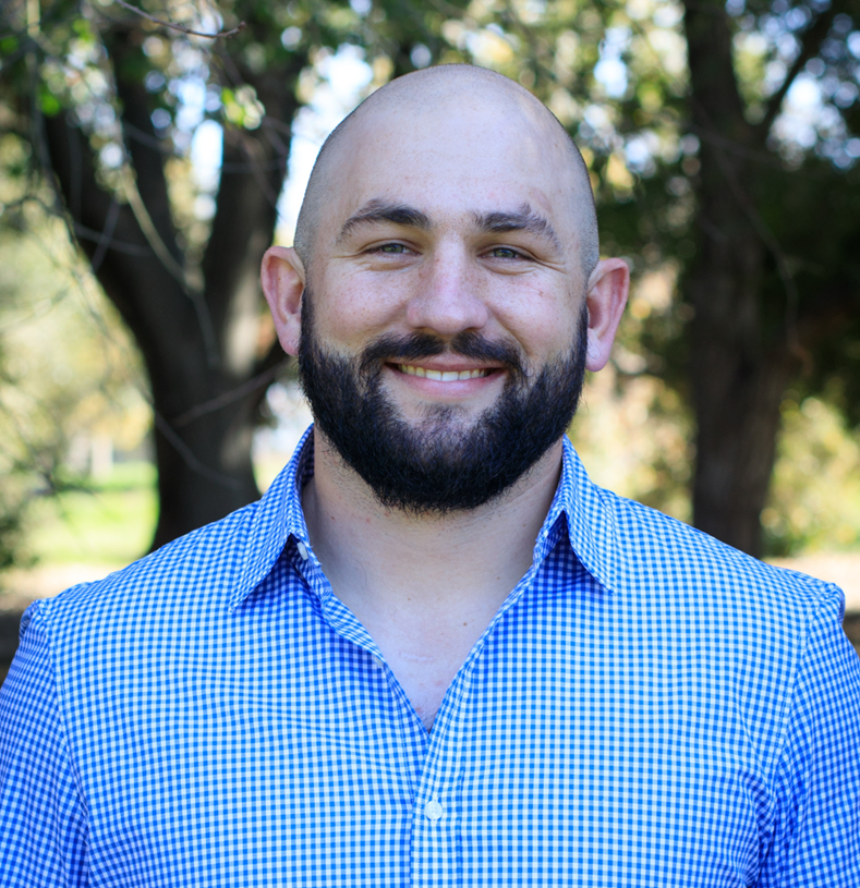 A person with a shaved head and a dark beard smiles while wearing a light blue checkered shirt, set against a blurred tree.
