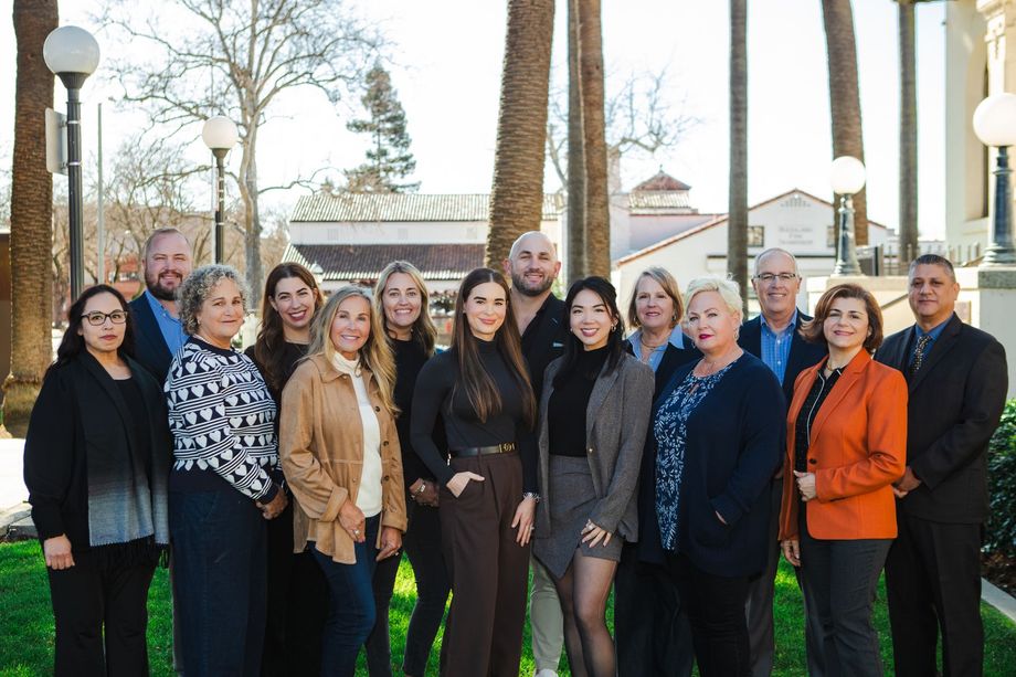 A group of people standing together for a photo in an outdoor, sunlit plaza with palm trees and a building in the background.