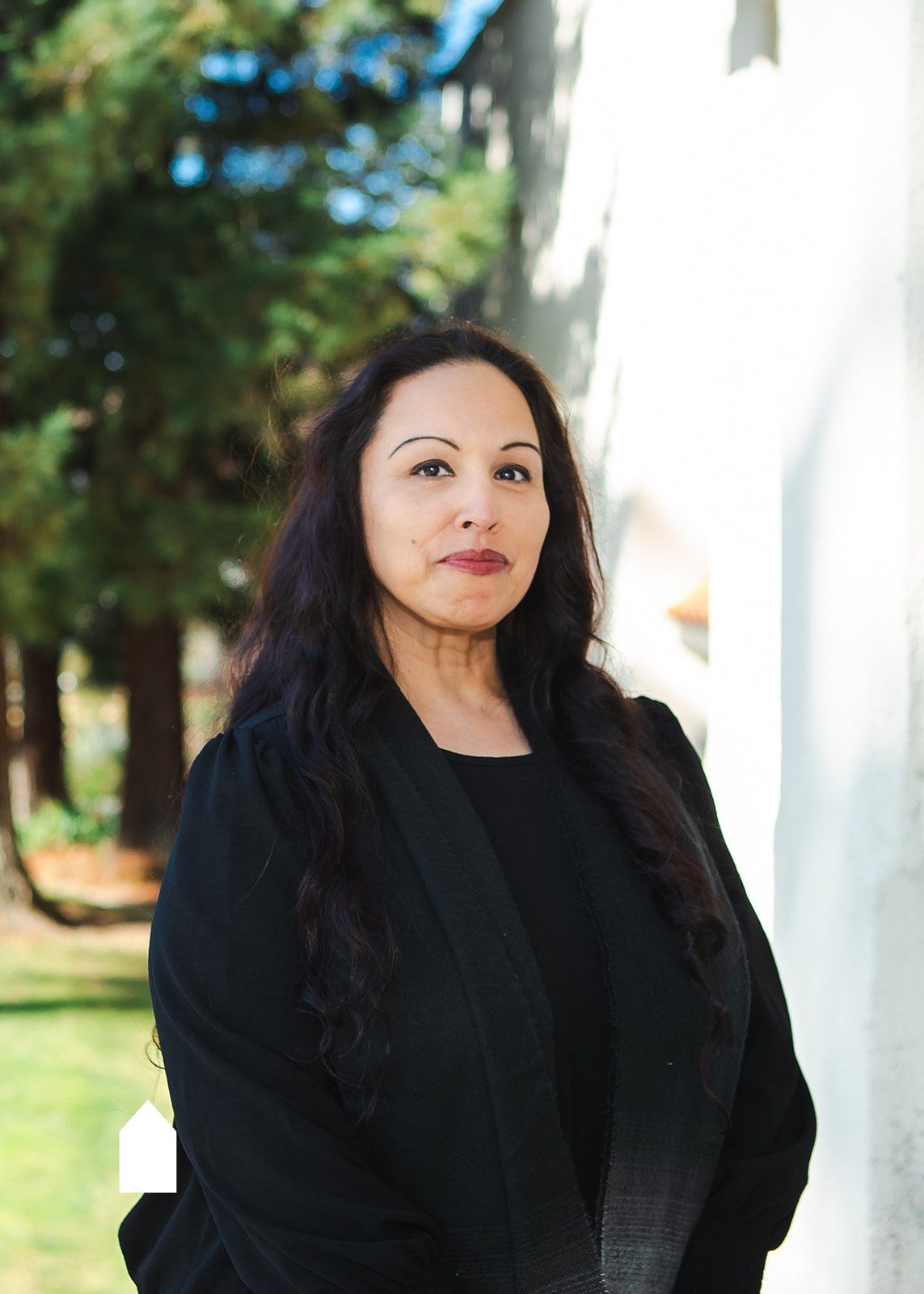 A person with long, dark hair wearing a black top and jacket, standing outdoors near a white wall and green trees.