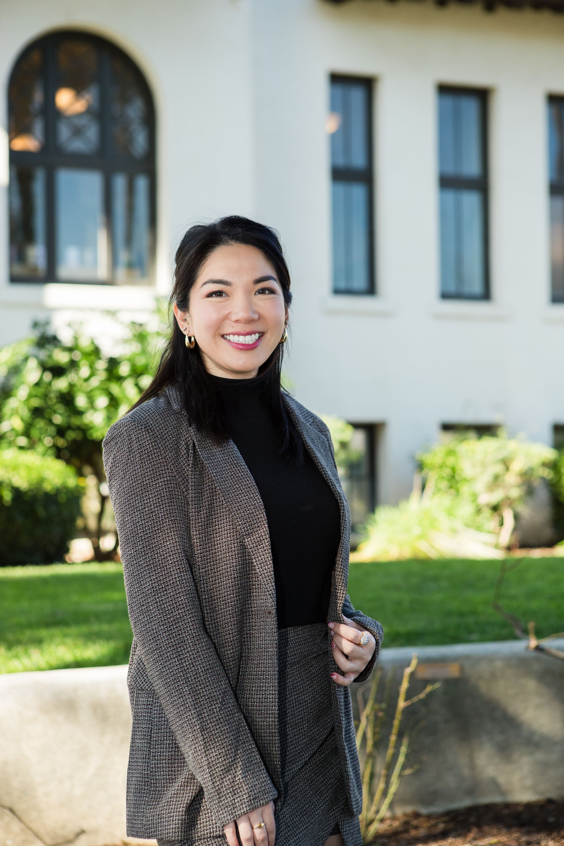 A smiling person in a patterned blazer and black turtleneck stands outdoors in front of a white building with windows.