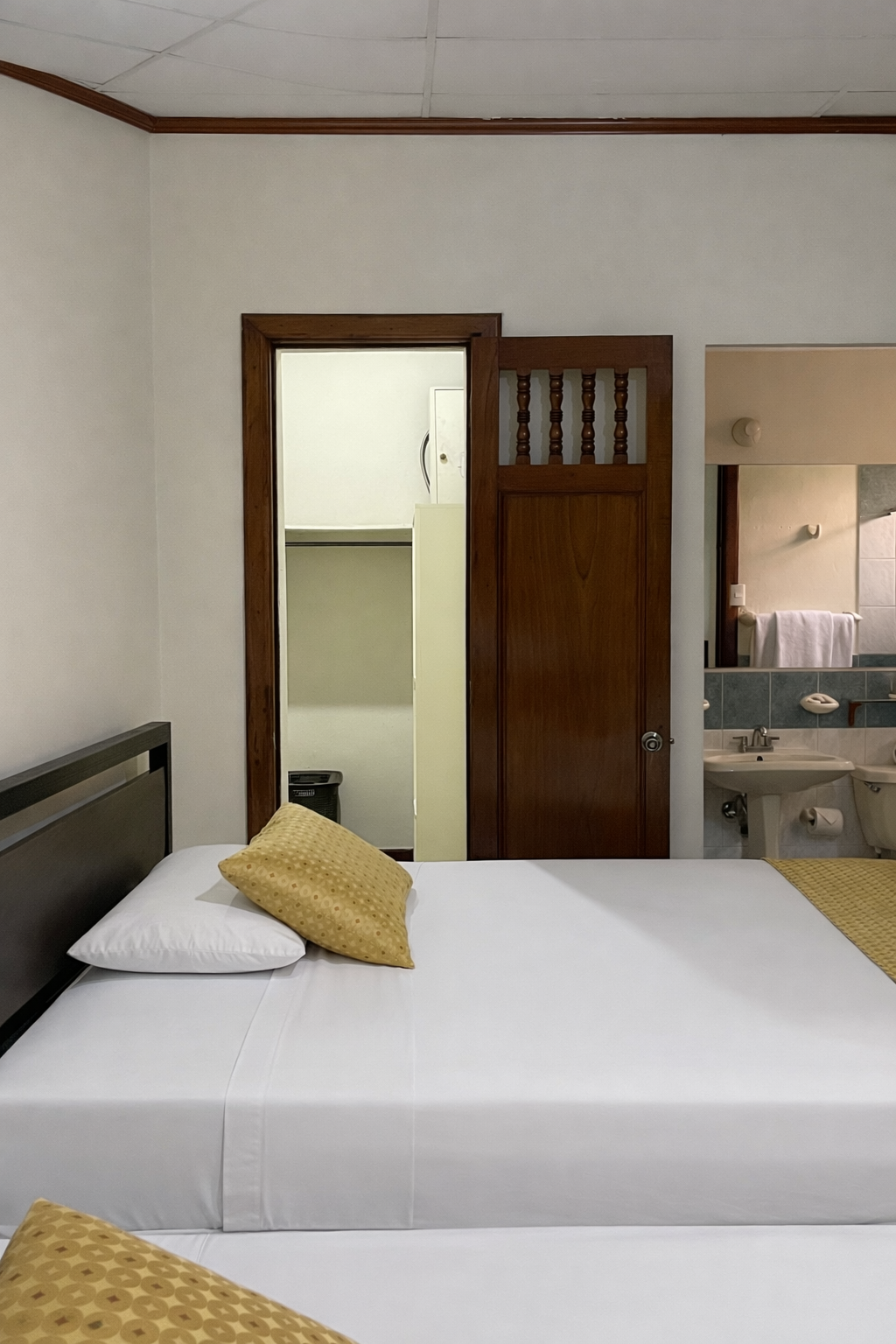 A hotel room with two beds, a wooden door leading to a bathroom, and a vanity area with a sink and a mirror.