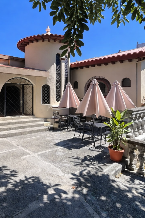 A sunny patio with beige stucco walls, terracotta roof tiles, and three umbrellas over outdoor seating.