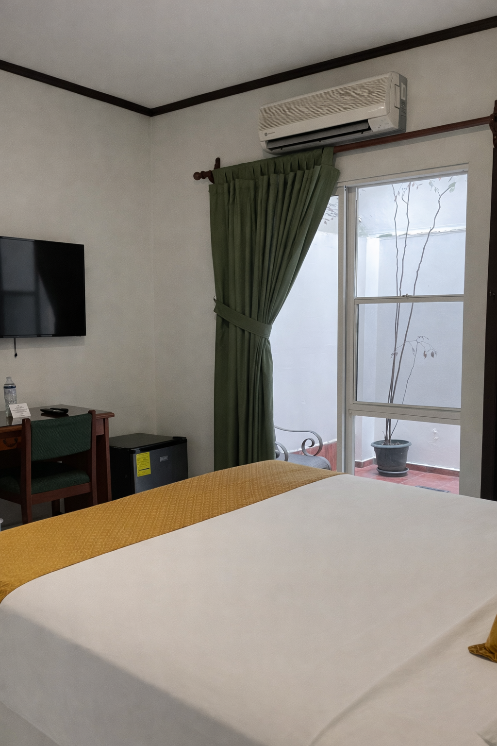 A hotel room with a bed featuring a gold runner, a desk with a chair, a wall-mounted TV, and a glass door with green drapes.