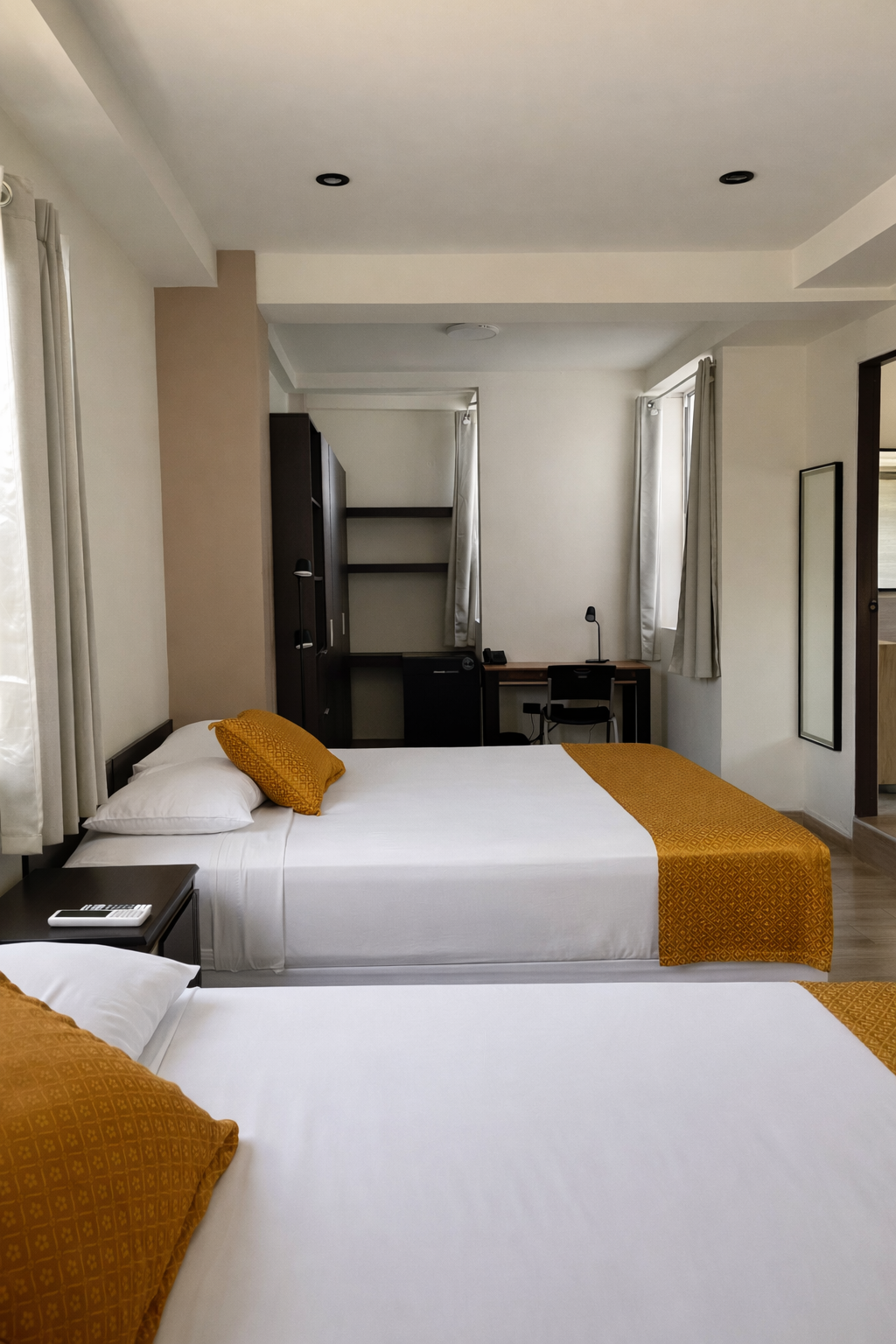 A hotel room with two white beds featuring gold runners, a desk, a tall wardrobe, and a mirror on the wall.