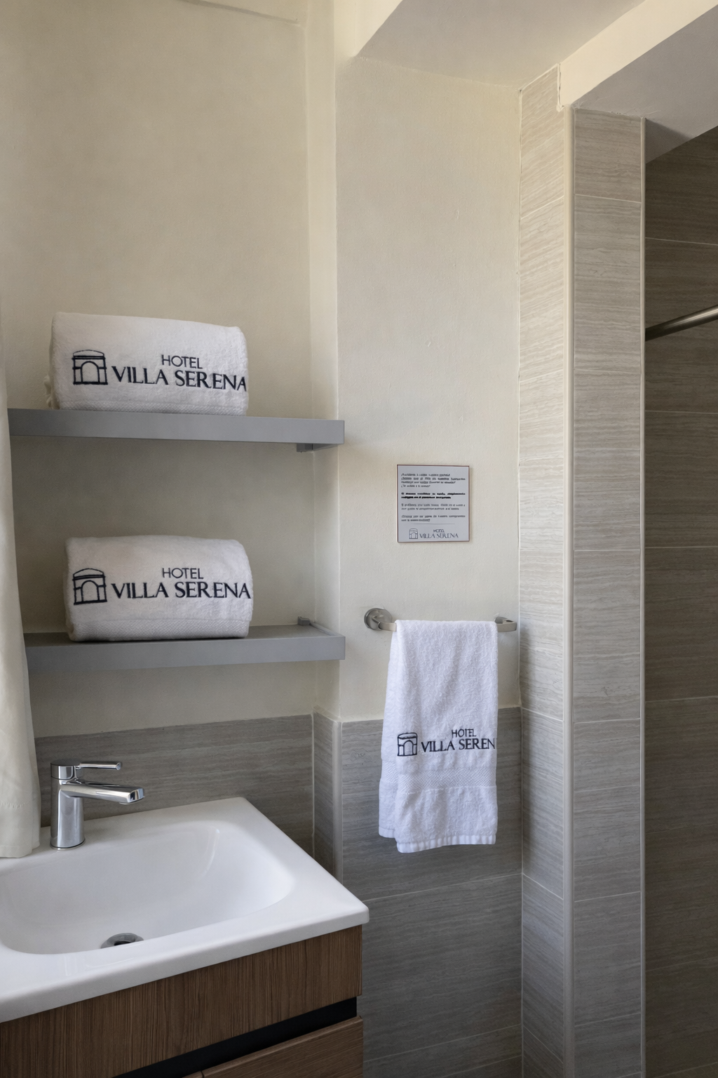 A hotel bathroom vanity with two gray shelves holding branded white towels, and a hand towel hanging beside a sink.
