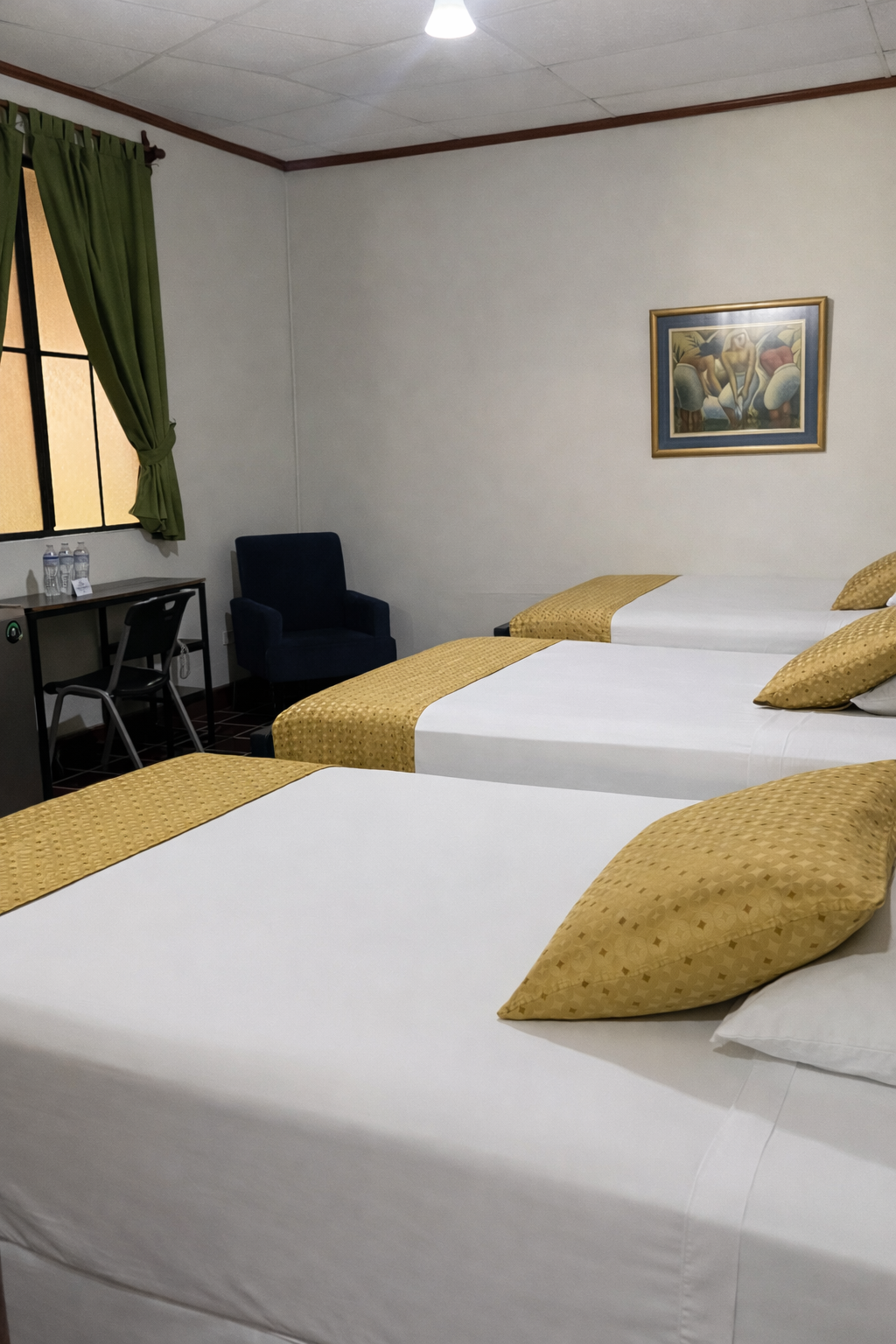 A hotel room featuring three beds with white linens and mustard yellow patterned runners, a desk, and a dark blue chair.