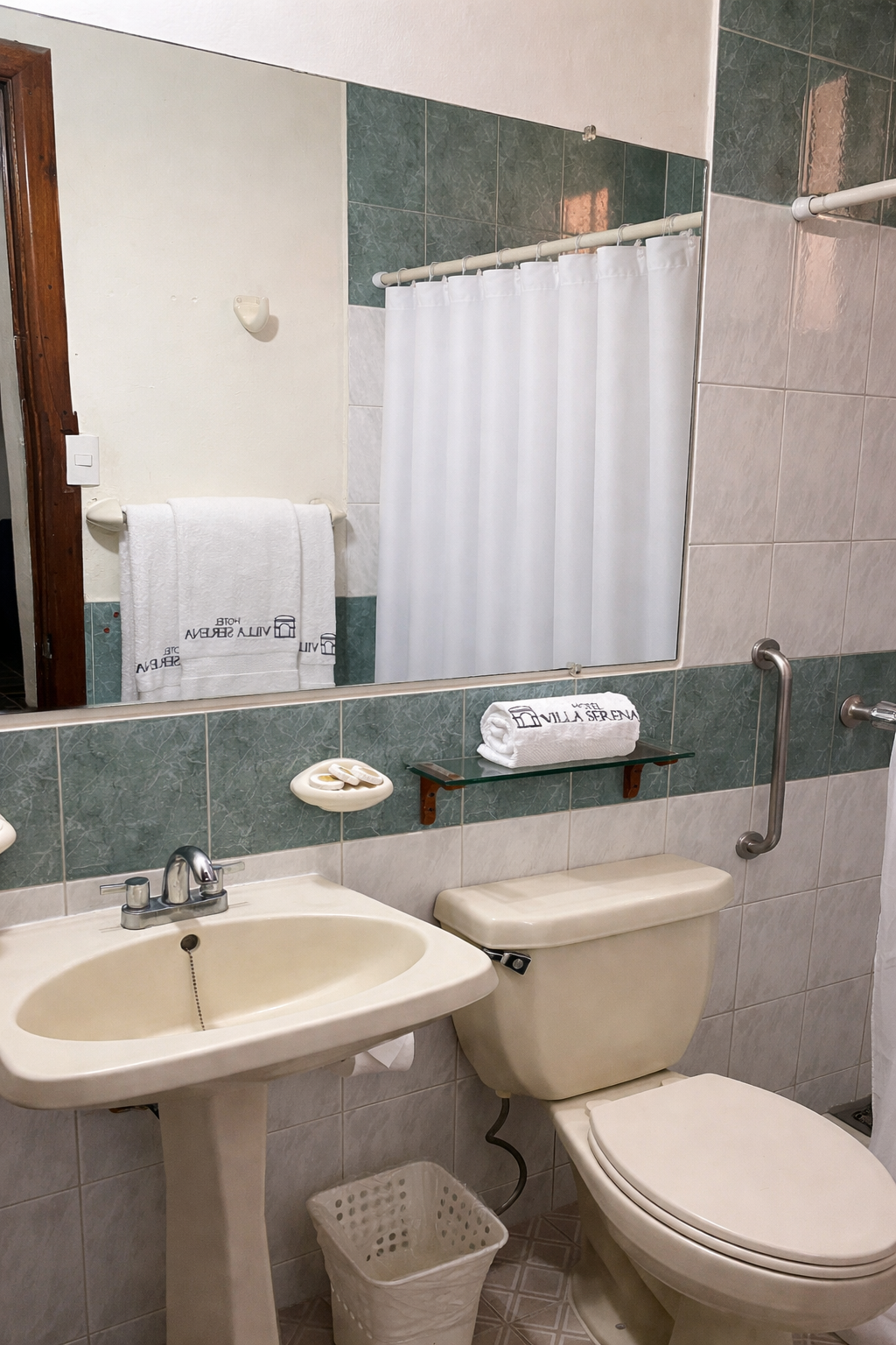 A hotel bathroom with a white pedestal sink, toilet, towel, and grab bar, featuring cream and green tiled walls.