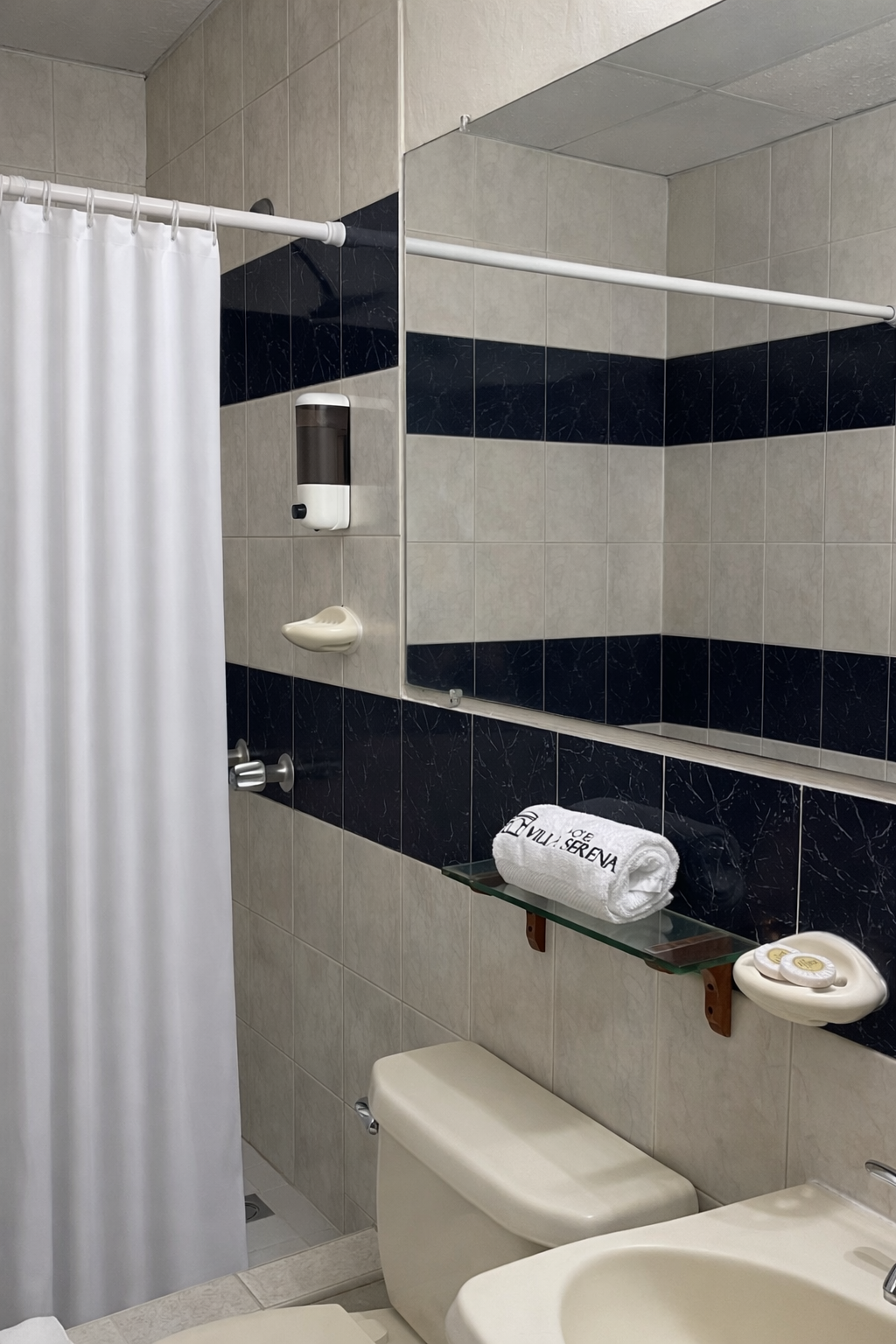 A hotel bathroom with a white shower curtain, a toilet, a sink, and a shelf with a rolled white towel and soap dish.