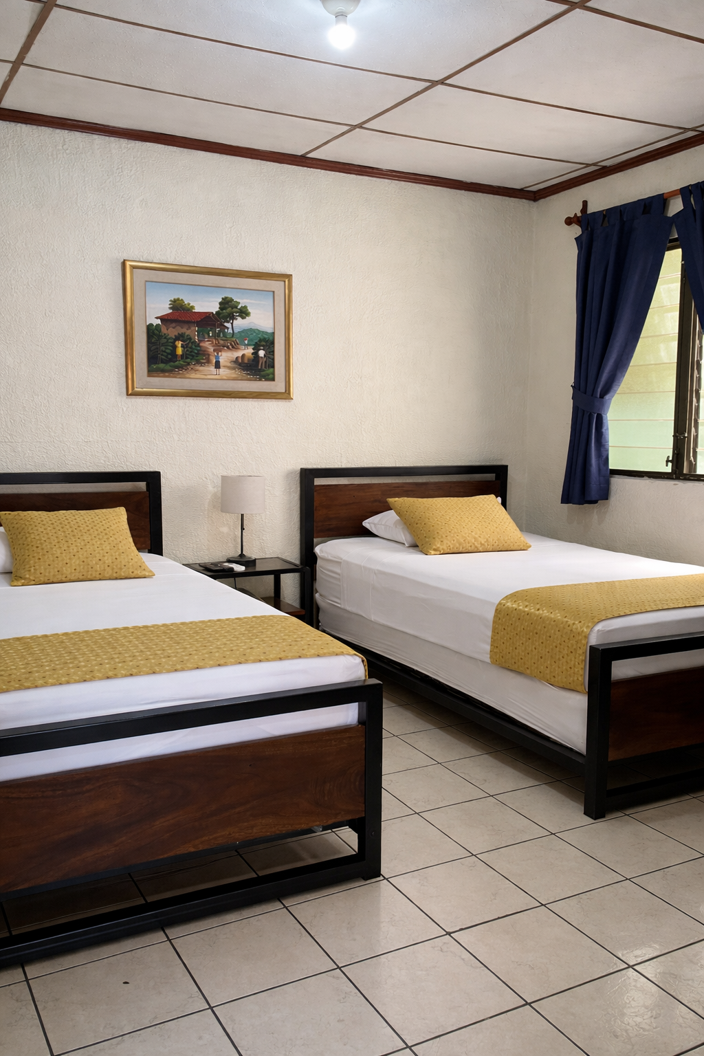 A hotel room with two dark wood beds, white bedding, gold runners, matching pillows, and blue curtains on a white wall.
