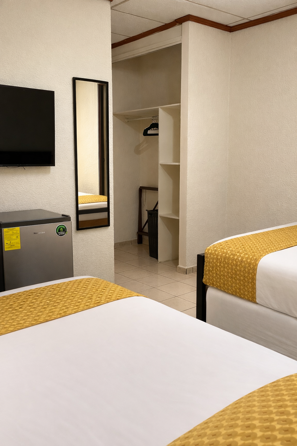 A hotel room with two beds, a small refrigerator topped by a TV, a wall mirror, and an open closet area.