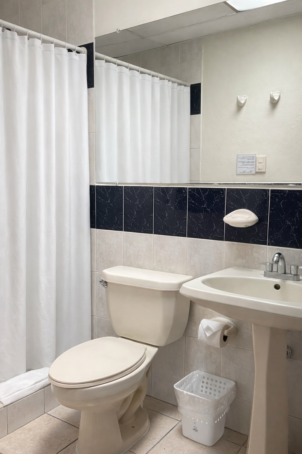 A small bathroom with a white toilet, pedestal sink, shower with a white curtain, and black tiled backsplash.