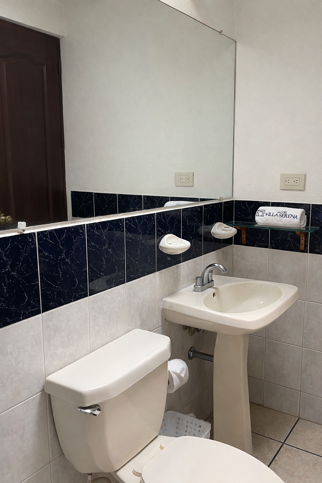 A bathroom with a white pedestal sink, a toilet, a mirror, and a wall featuring dark blue and white square tiles.