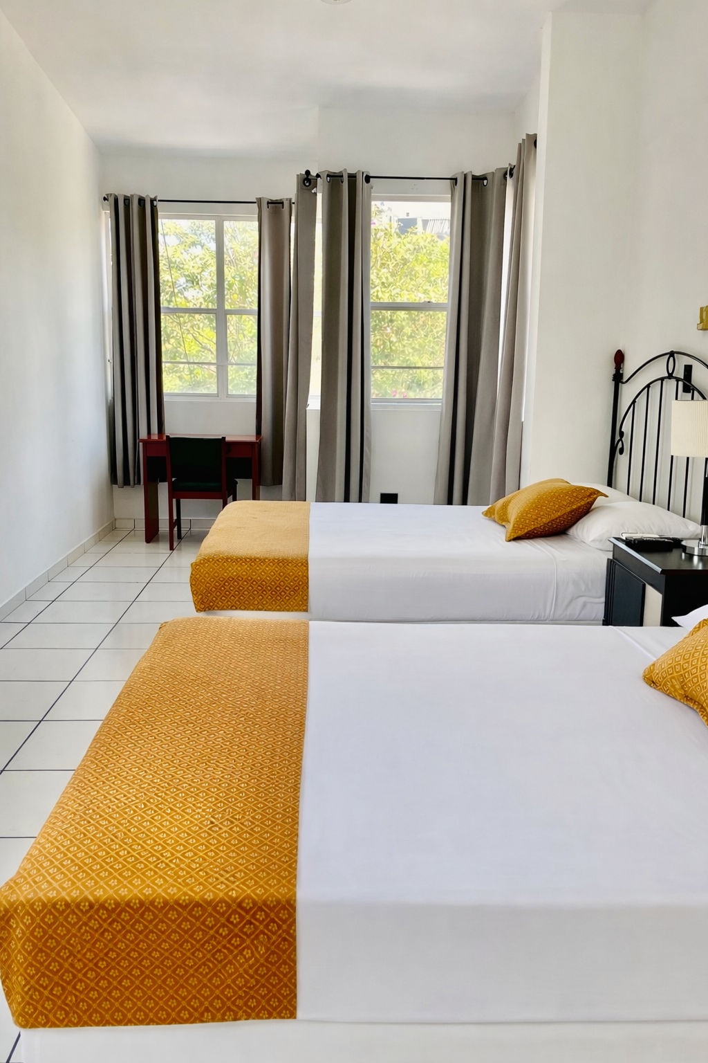 A hotel room with two beds featuring white linens and yellow runners, positioned near a window with floor-length curtains.