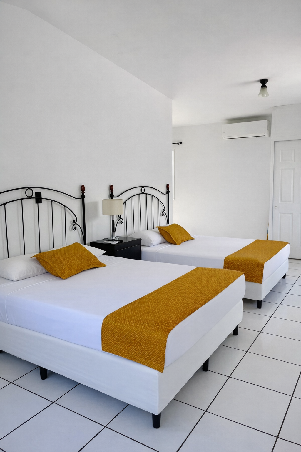 A bright hotel room with two white beds featuring black iron headboards and mustard-yellow runners on white tiled floors.