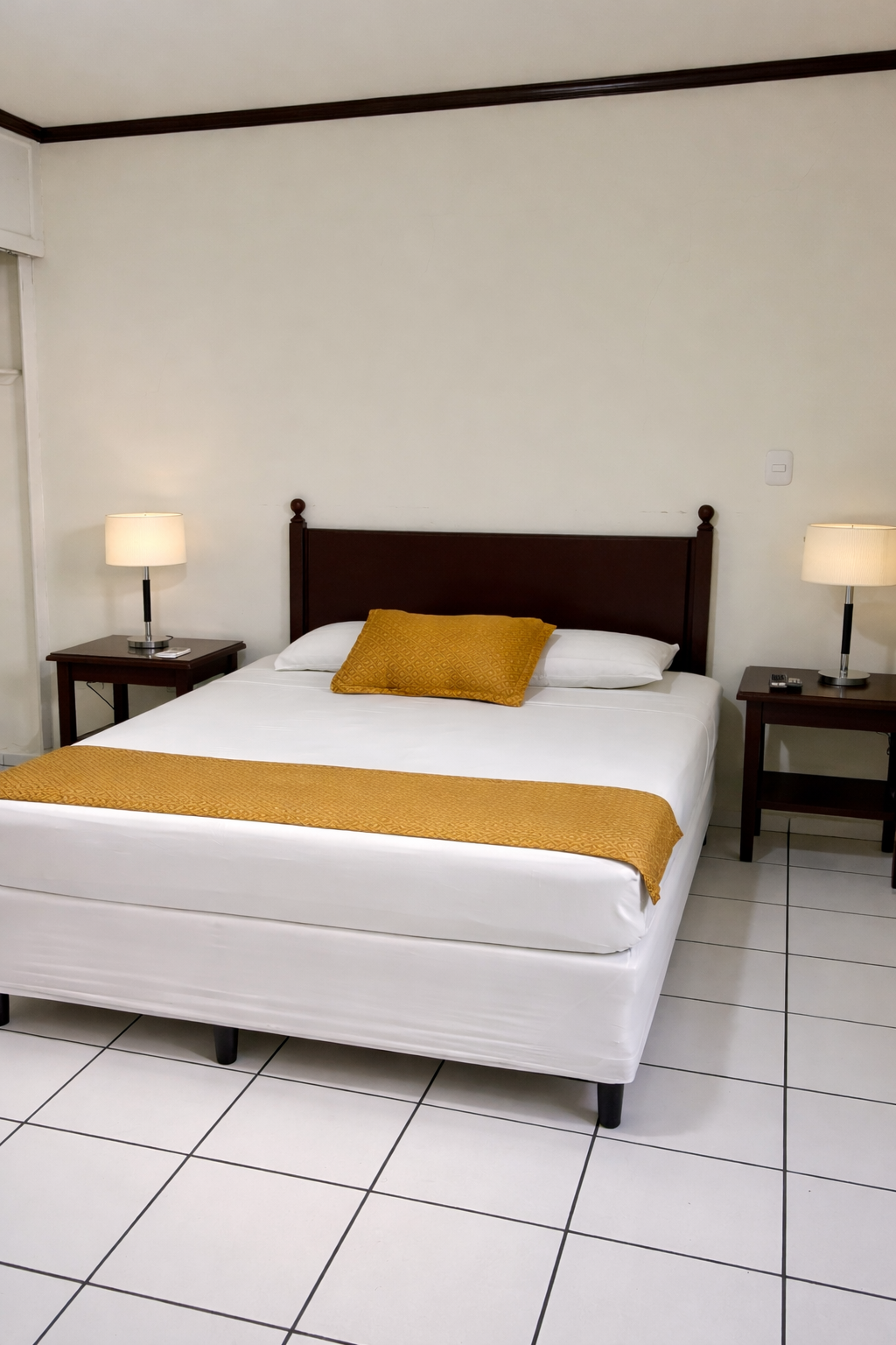 A made bed with white sheets and a yellow runner and pillow, flanked by two wooden nightstands with lamps, on white tile.