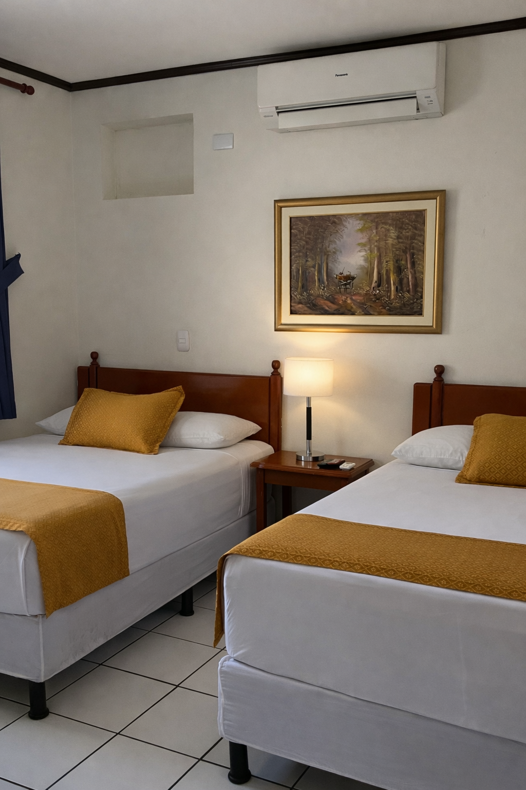 A hotel room with two beds featuring white linens, gold runners, and matching accent pillows, under a wall-mounted AC unit.