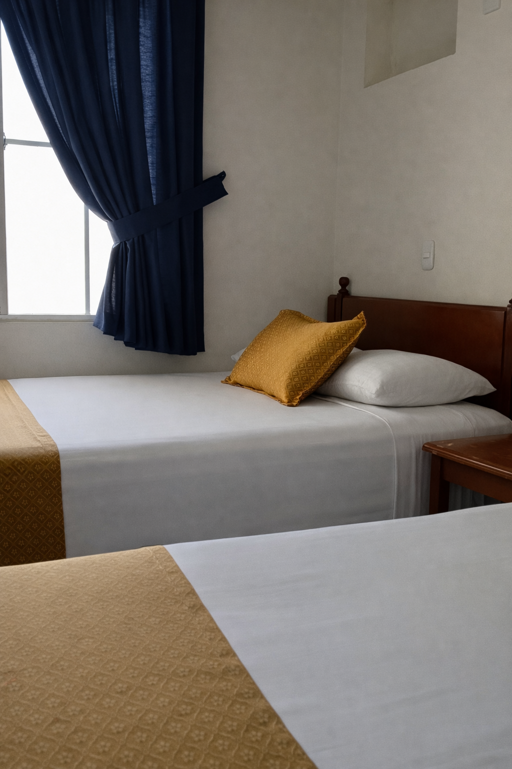 A hotel room with two beds, featuring white linens with gold runners, dark blue curtains, and wood headboards.