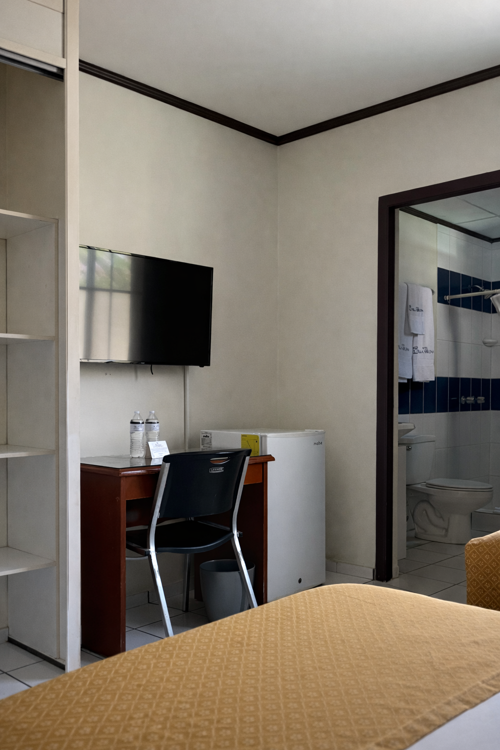 A hotel room featuring a television mounted on the wall, a wooden desk with a chair, a mini-fridge, and an open bathroom.