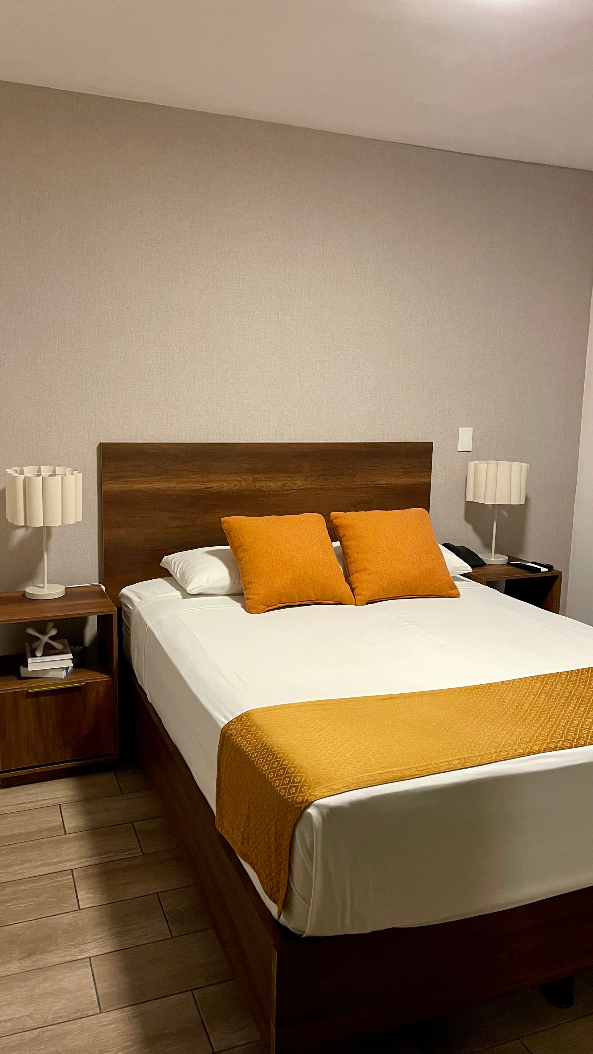 A hotel-style bedroom with a bed featuring a wooden headboard, white bedding, and orange accent pillows and runner.