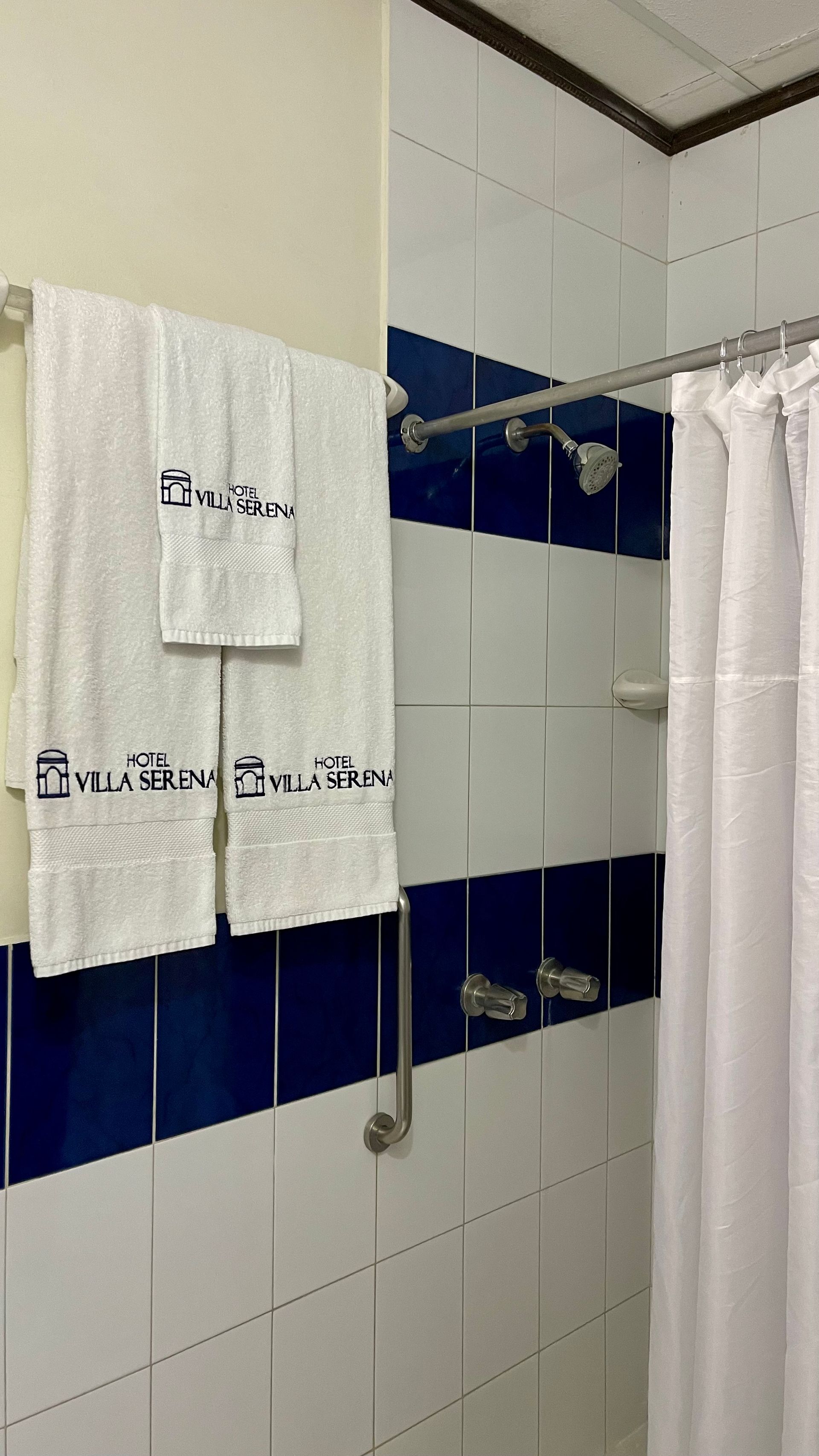 A hotel bathroom shower area with white and blue tiled walls, white towels on a rack, and a white shower curtain.