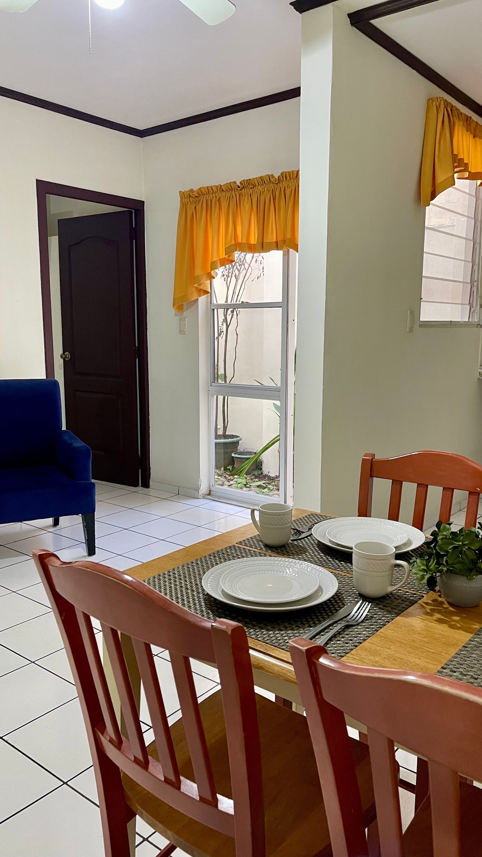 A dining table with two wooden chairs, place settings, and a plant, near a blue armchair and a door with yellow curtains.