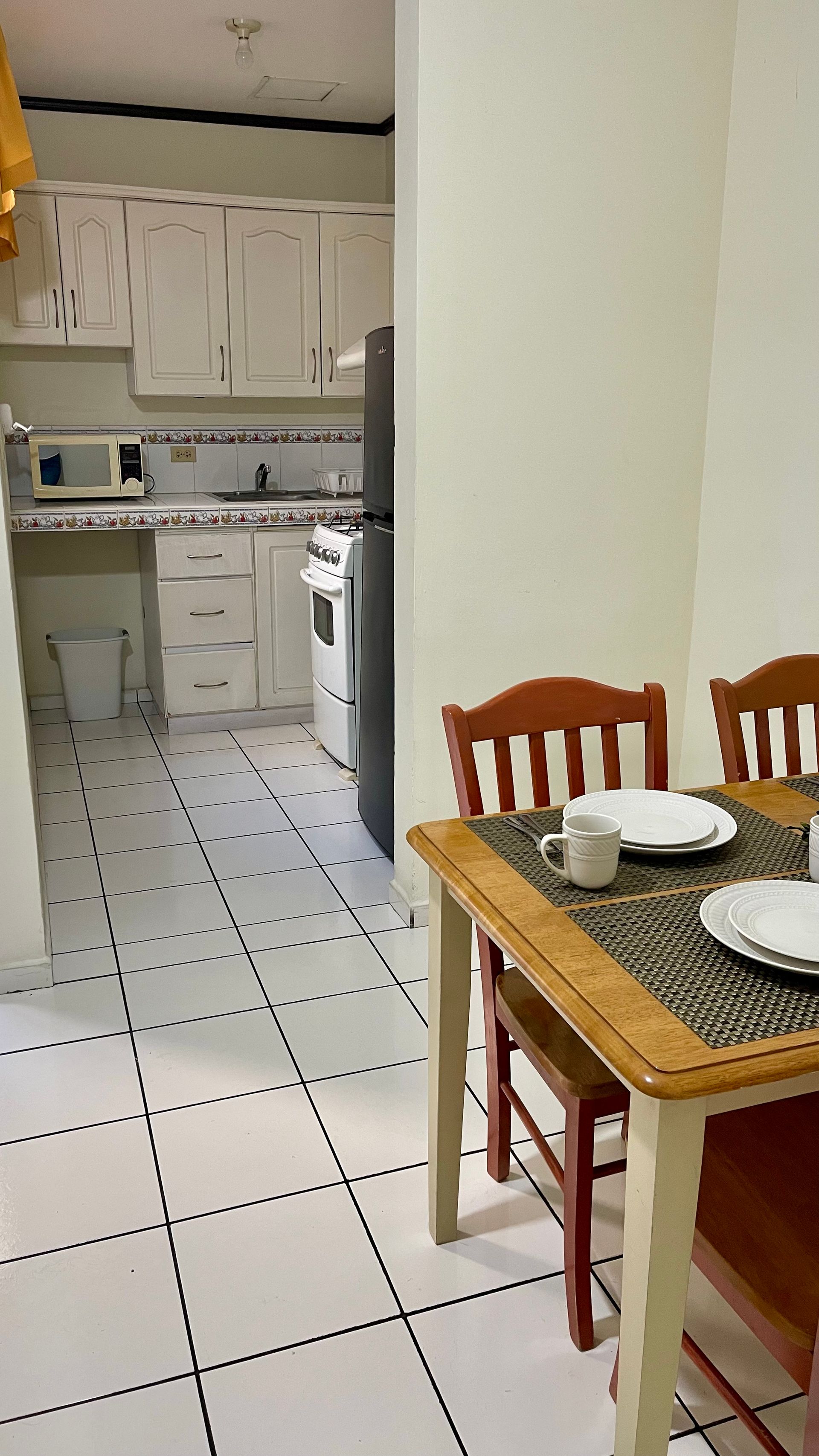 A dining table with place settings in the foreground, looking into a white kitchen with tiled floors and a refrigerator.