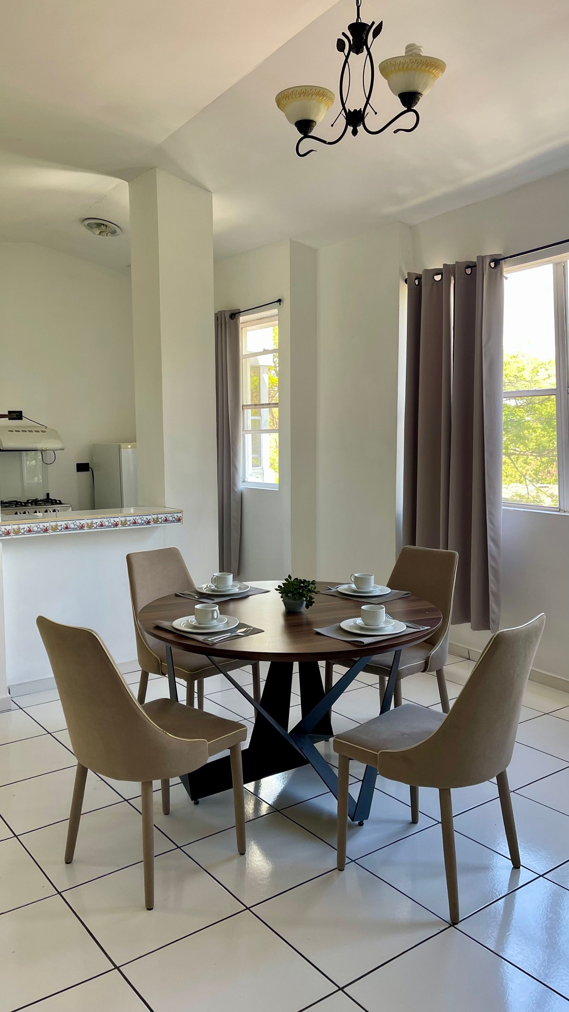 A dining area with a round wooden table, four tan chairs, tile floors, and a pendant light, next to a kitchen counter.