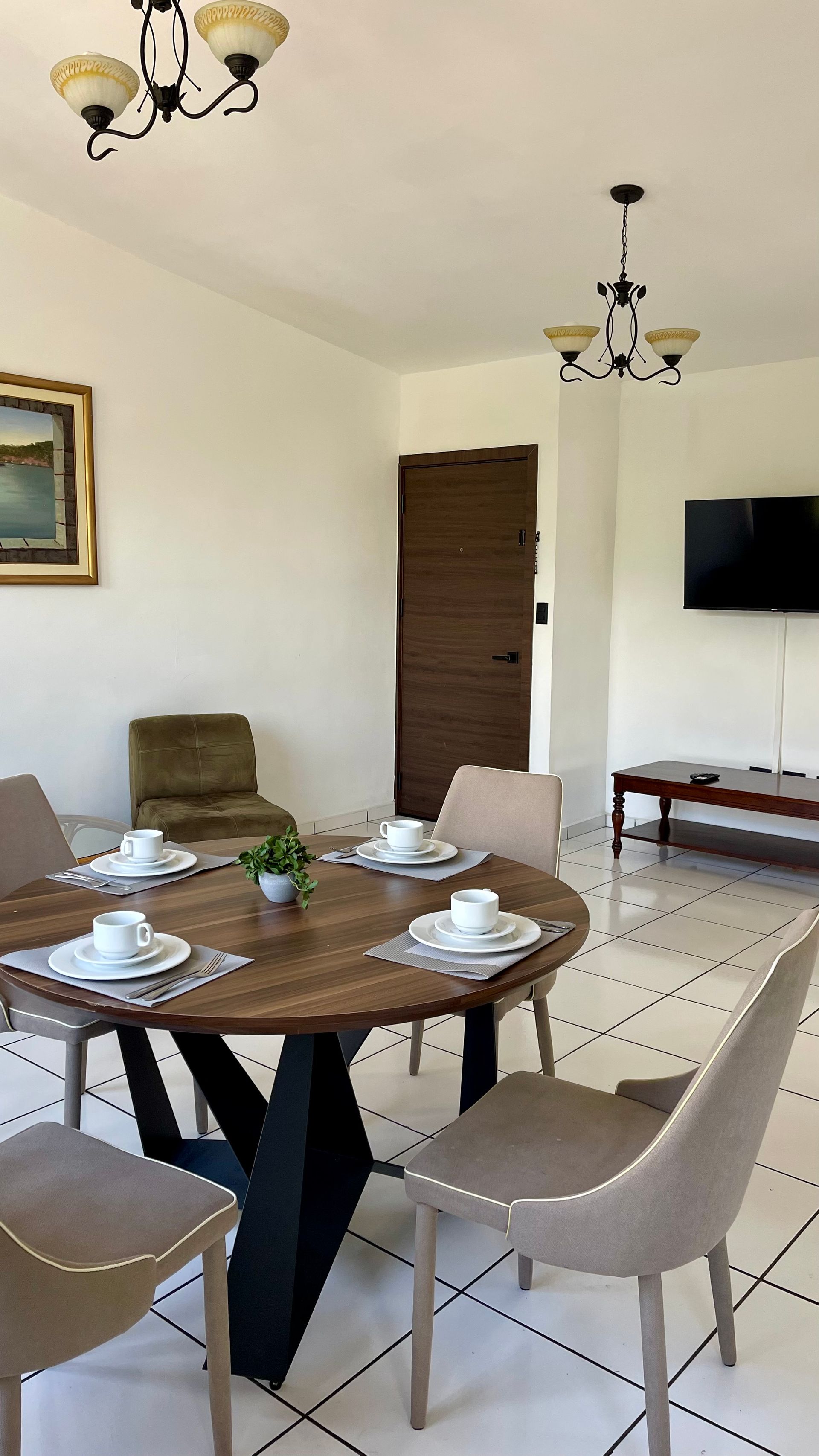 A dining area featuring a round wood table with four beige chairs, two overhead lamps, a painting, and a wall-mounted TV.