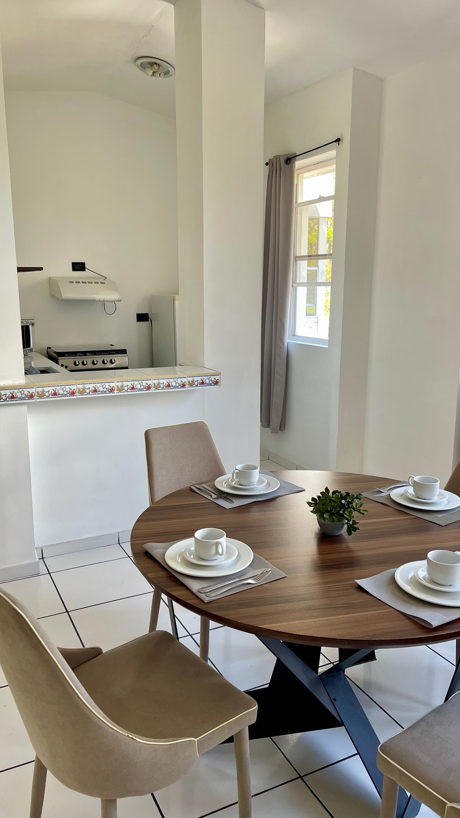 A circular wooden dining table set for four with white plates and mugs, located in a bright dining room with tile floors.