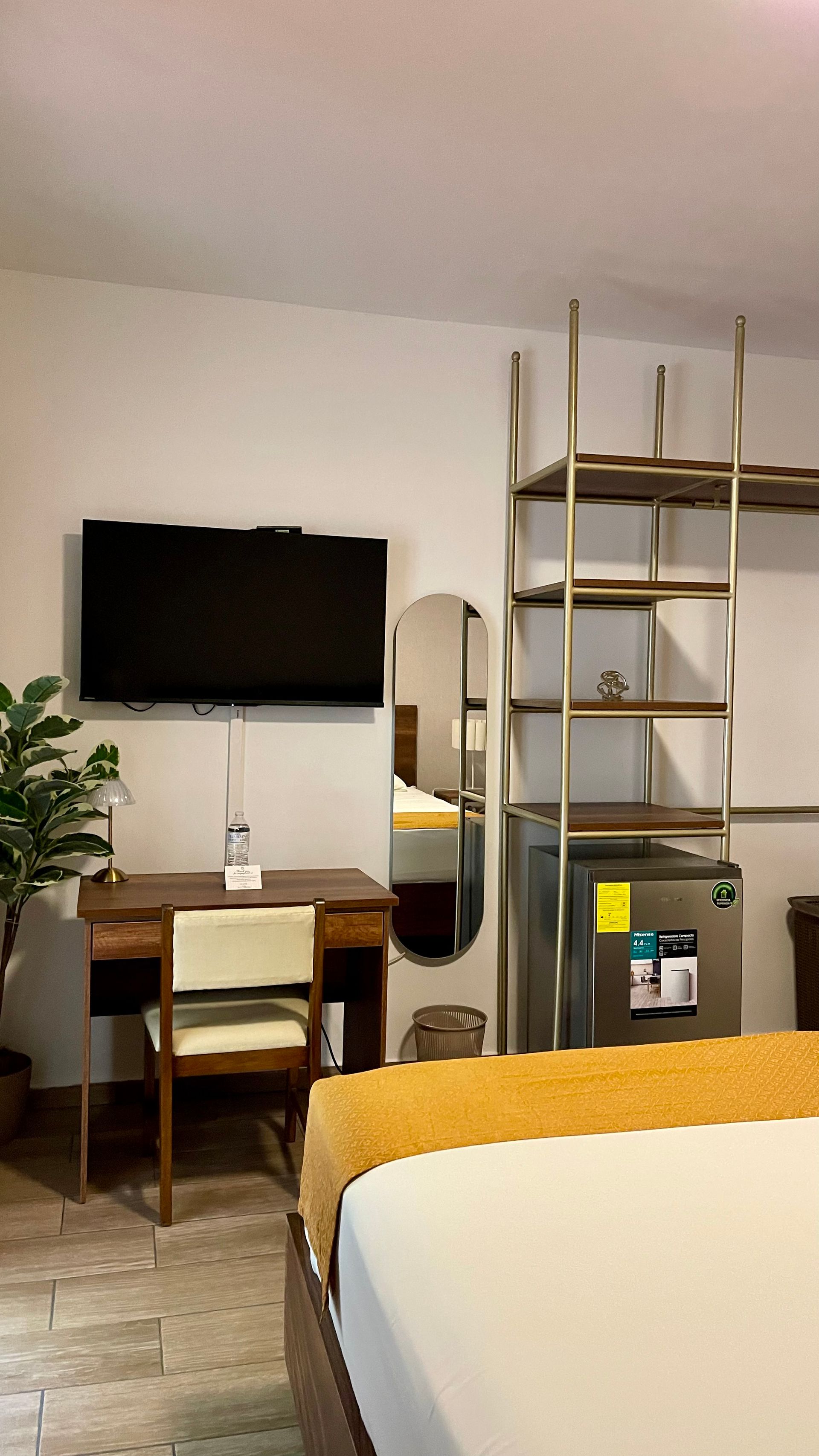 A hotel room with a desk, a mounted television, a floor mirror, an open metal shelving unit, and a small refrigerator.
