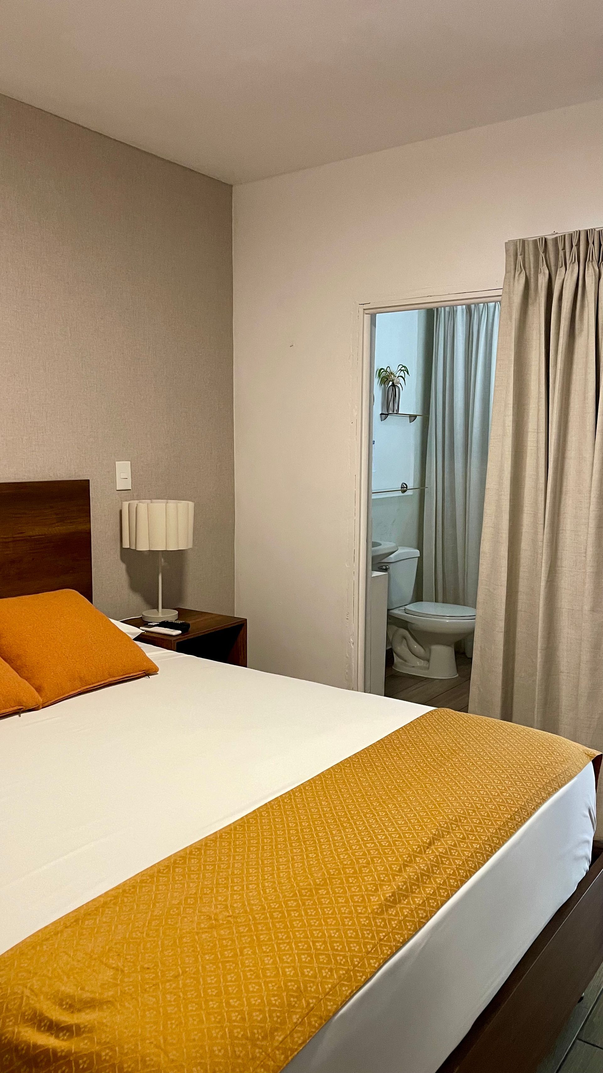A hotel room featuring a bed with a white sheet, orange pillows, and a yellow runner, with a bathroom visible in the back.