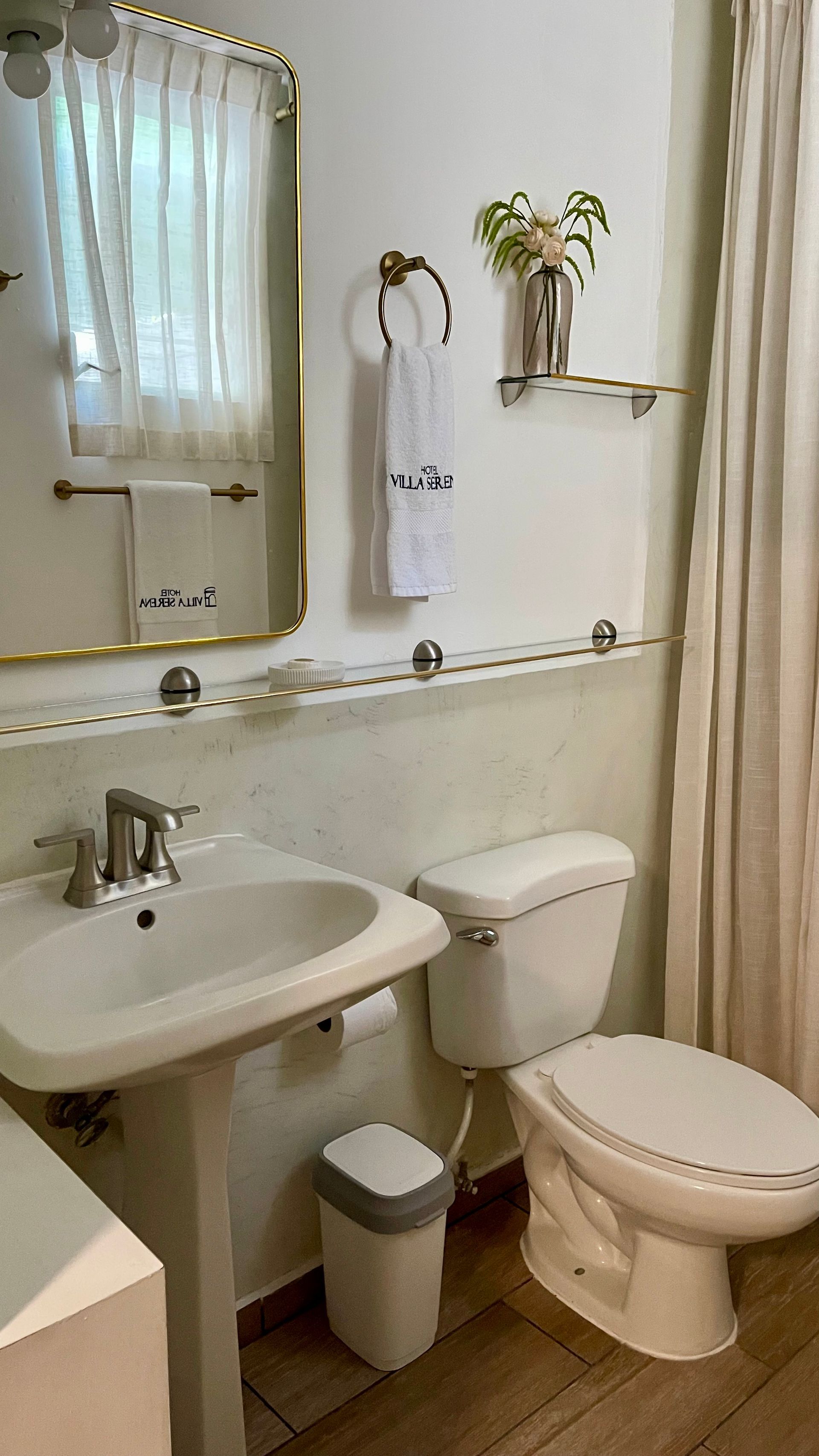 A clean bathroom with a pedestal sink, gold-rimmed mirror, white toilet, small trash bin, and decor on a small shelf.