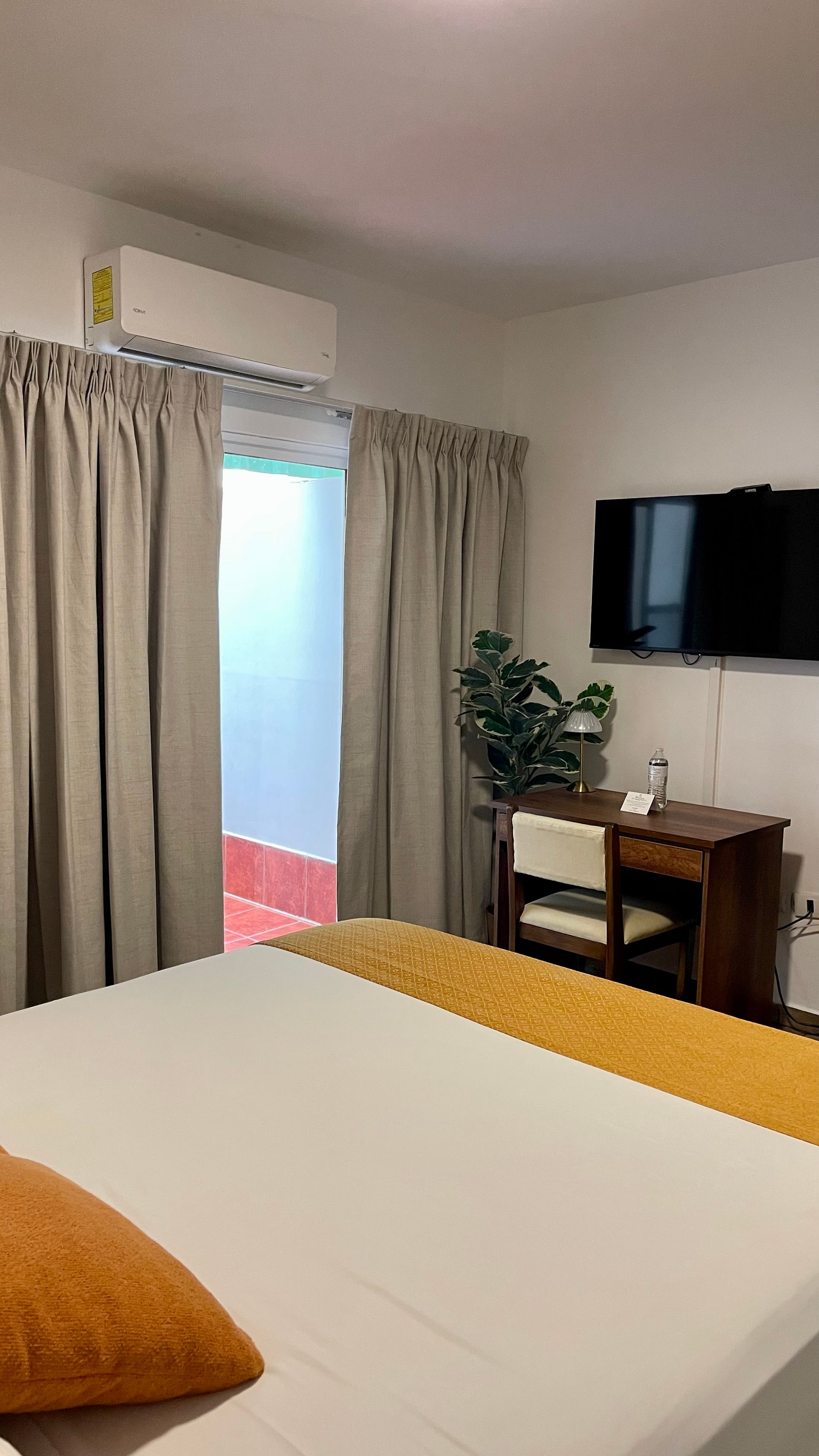 A neutral-toned bedroom with a bed featuring a gold runner, a small wooden desk, a television, and floor-length curtains.