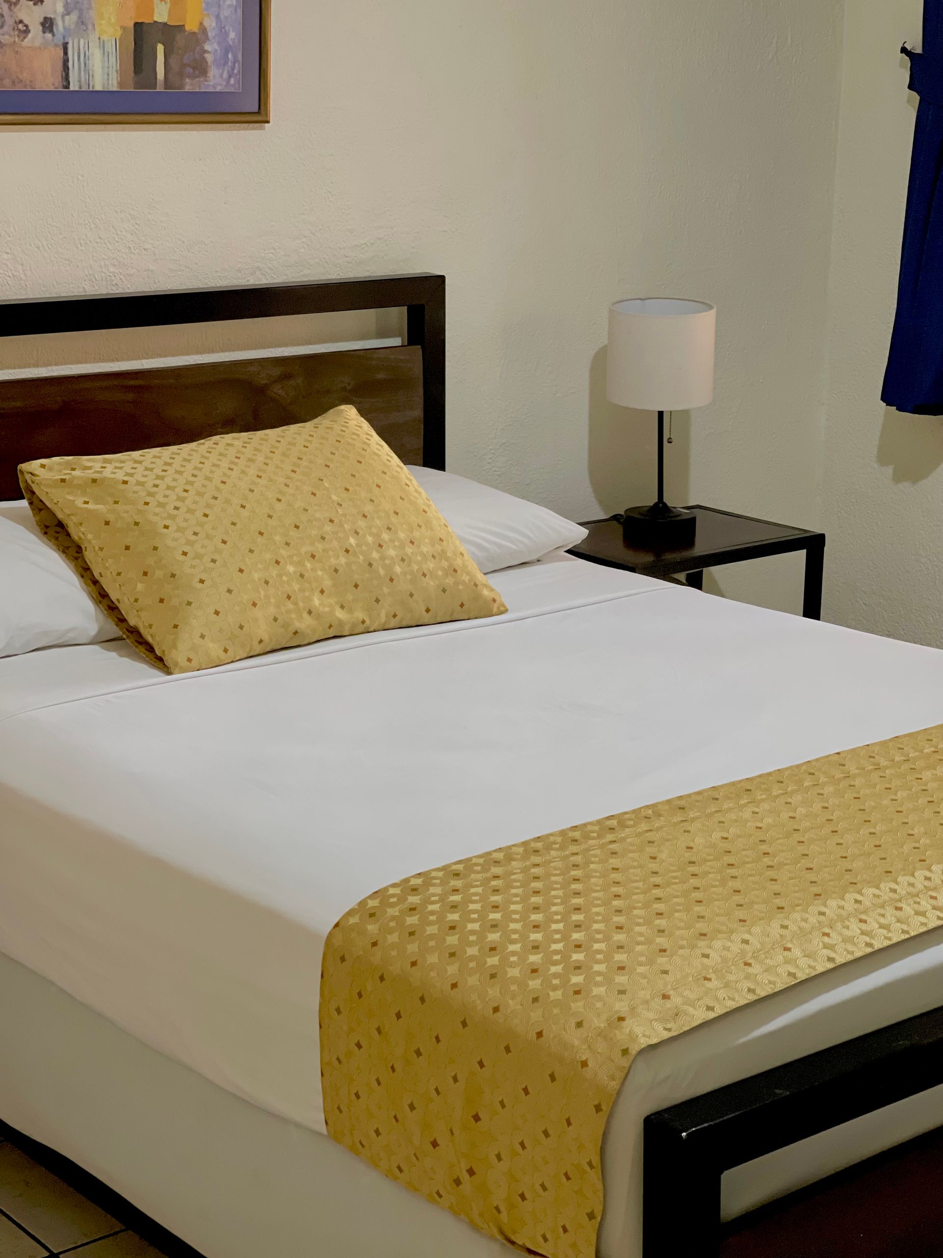 A neatly made bed with white sheets, a gold patterned runner and pillow, and a lamp on a bedside table.