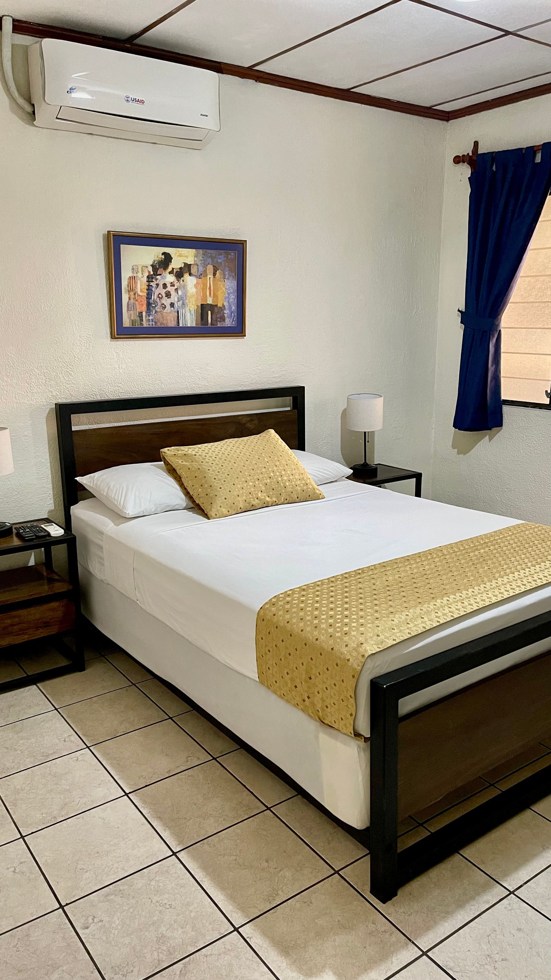 A furnished bedroom with a bed featuring a dark frame, white bedding, and a yellow decorative runner, with wall art above.