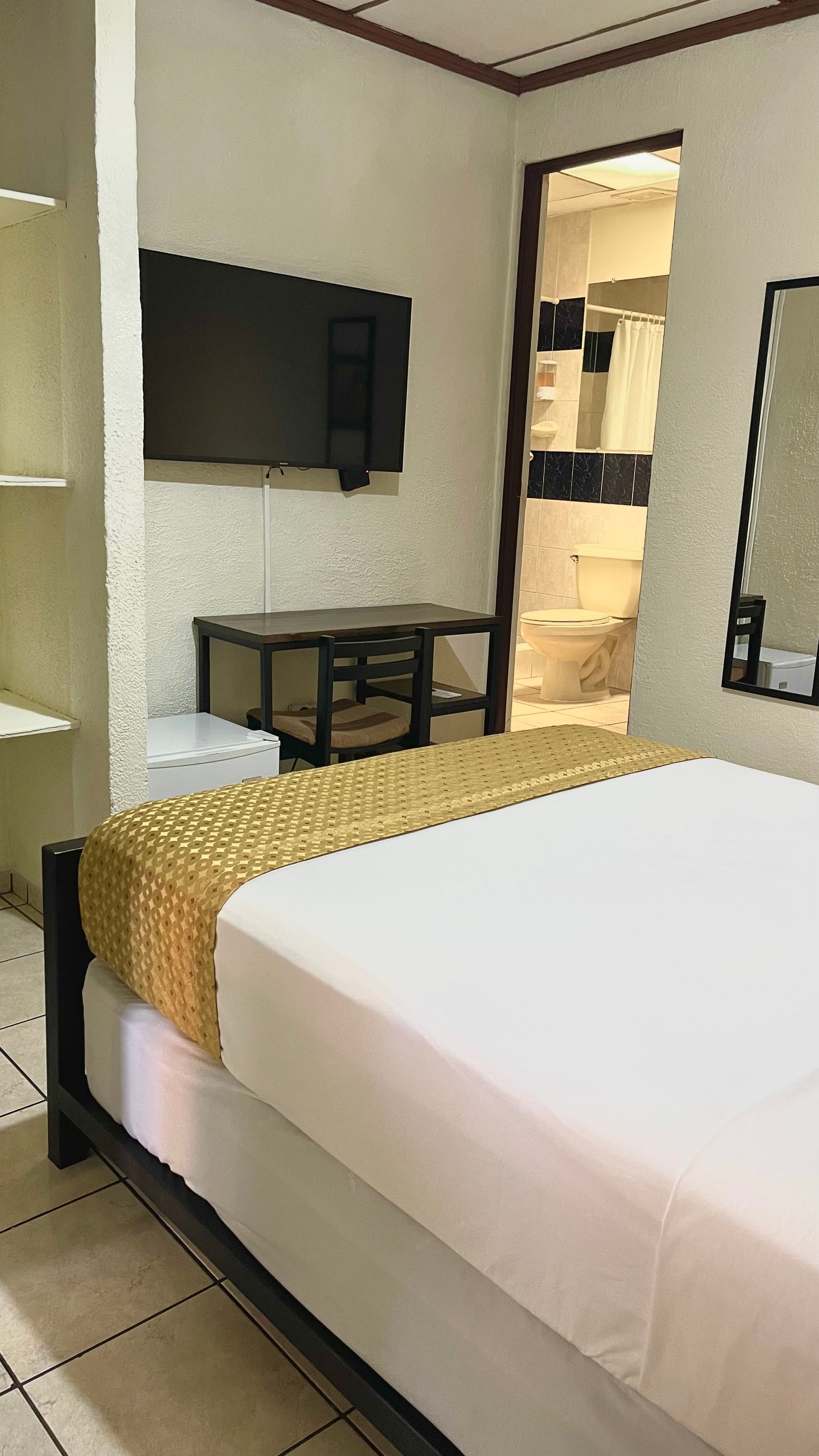 A hotel room with a queen bed with white linens and a gold runner, a TV, a small desk, and a view into the bathroom.